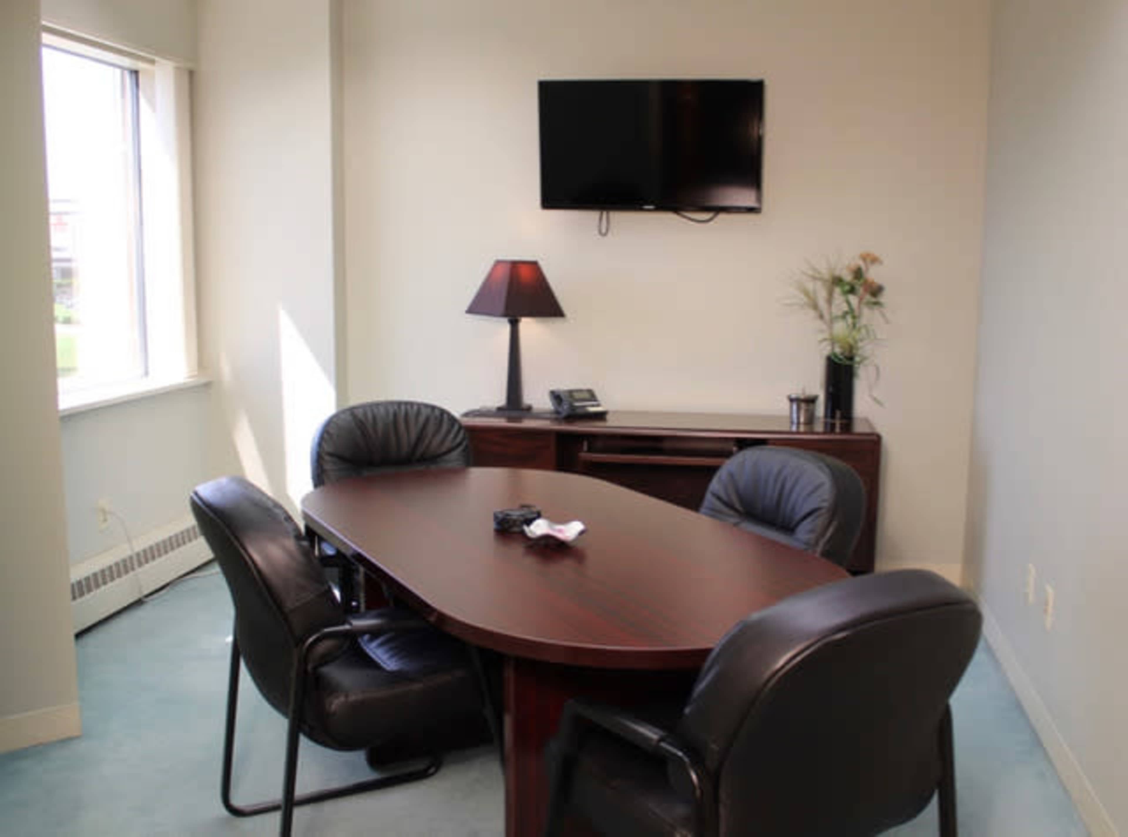 The image shows a small conference room with a wooden table, four black chairs, a wall-mounted television, and a lamp on a side table.