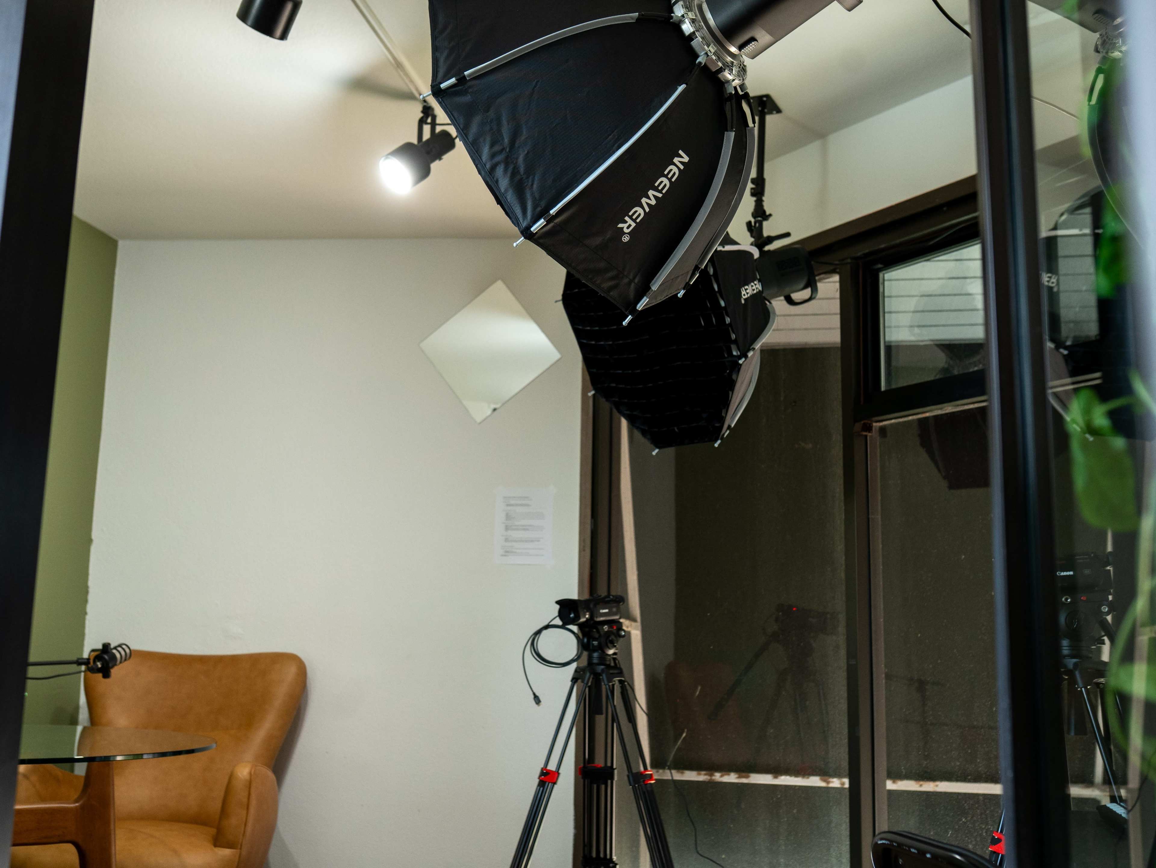 The image shows a photography studio setup with softbox lights, a camera on a tripod, and a brown armchair near a glass table.