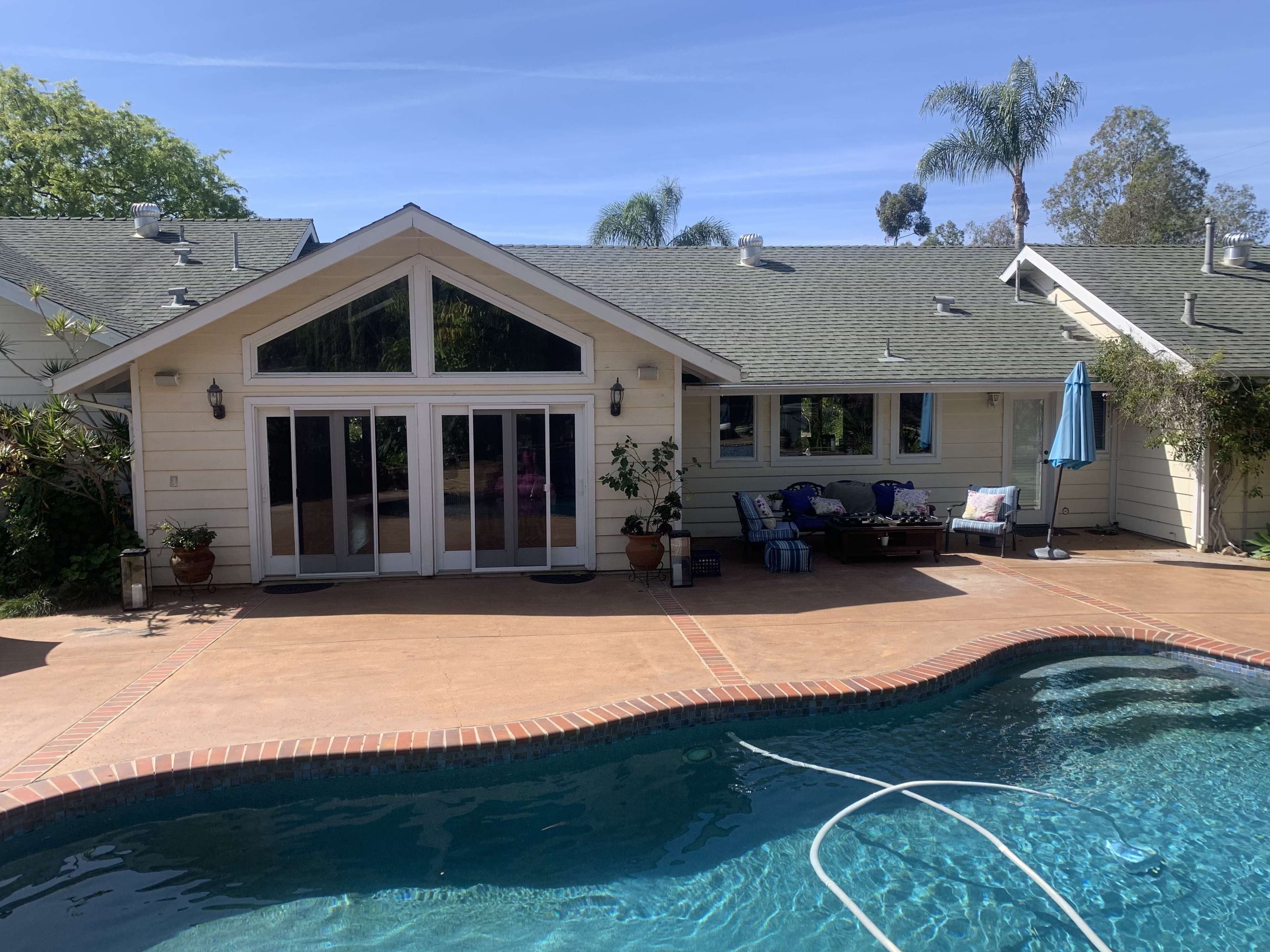 A backyard with a swimming pool in front of a house featuring large glass doors and a green lawn surrounded by palm trees.