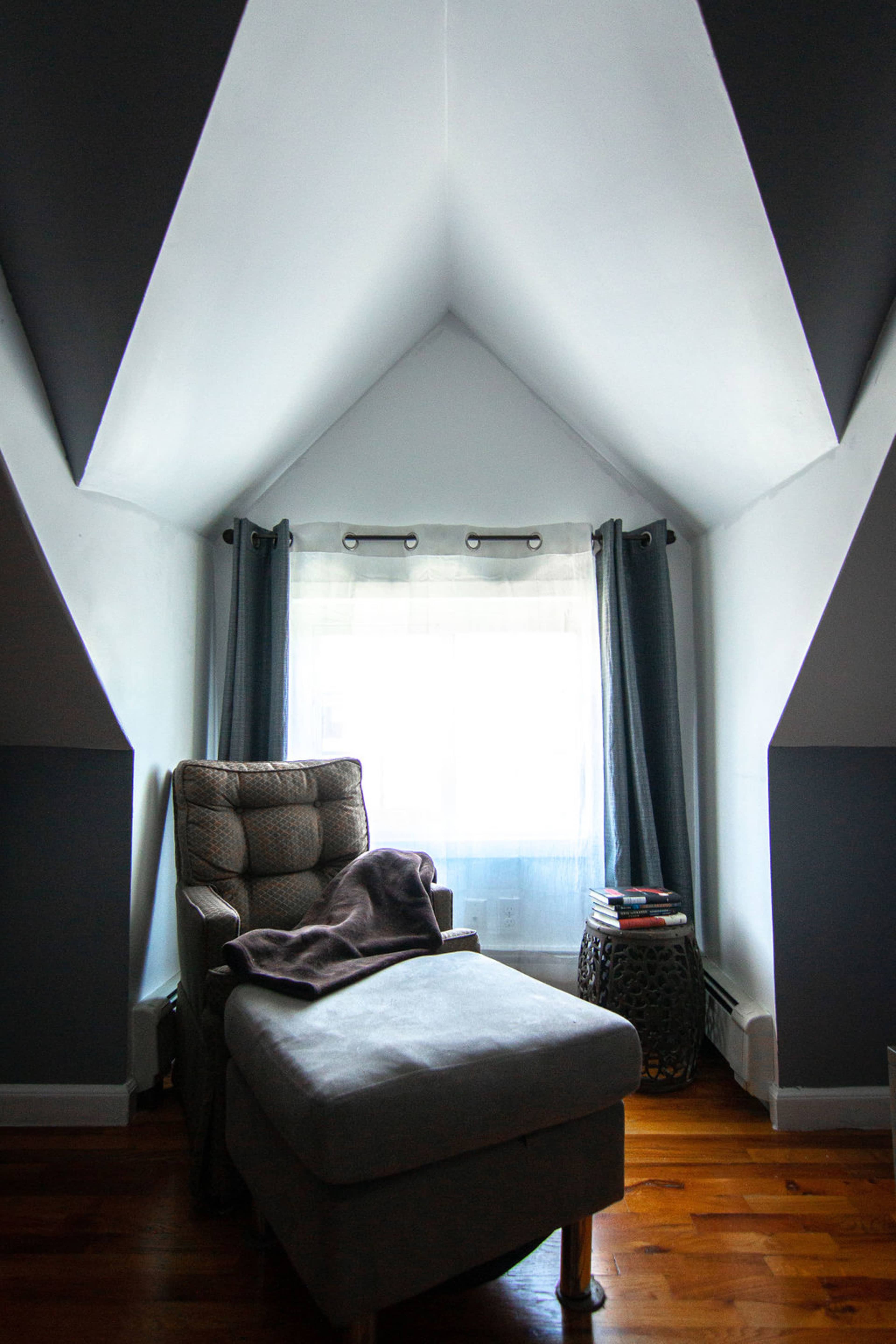A cozy reading nook features a gray chaise lounge next to a window, framed by curtains and a small side table.