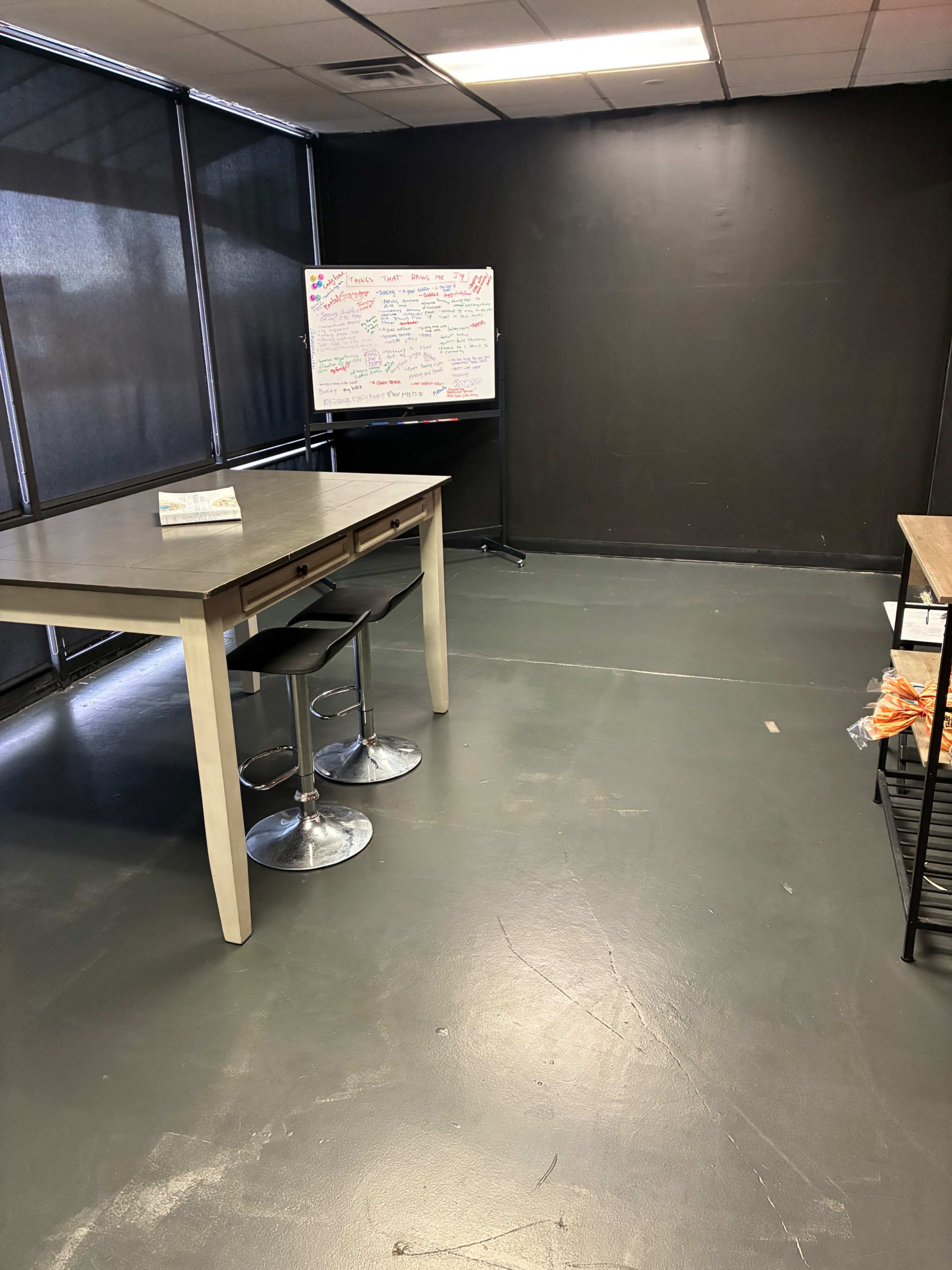 The image shows a sparsely furnished room with a table and two stools, a whiteboard filled with notes in the background, and dark walls.