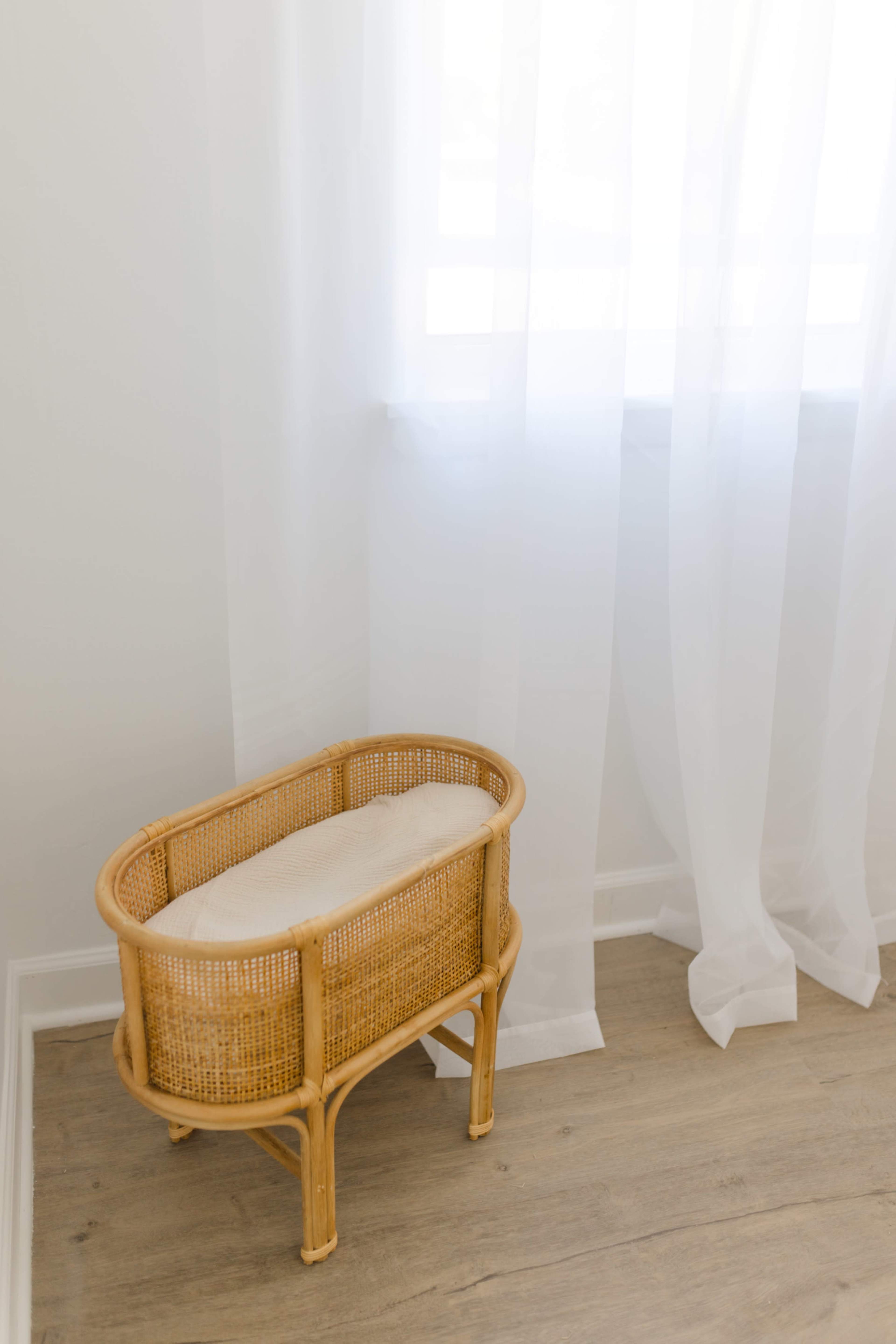A small wicker bassinet with a white mattress sits beside a window covered with sheer curtains.