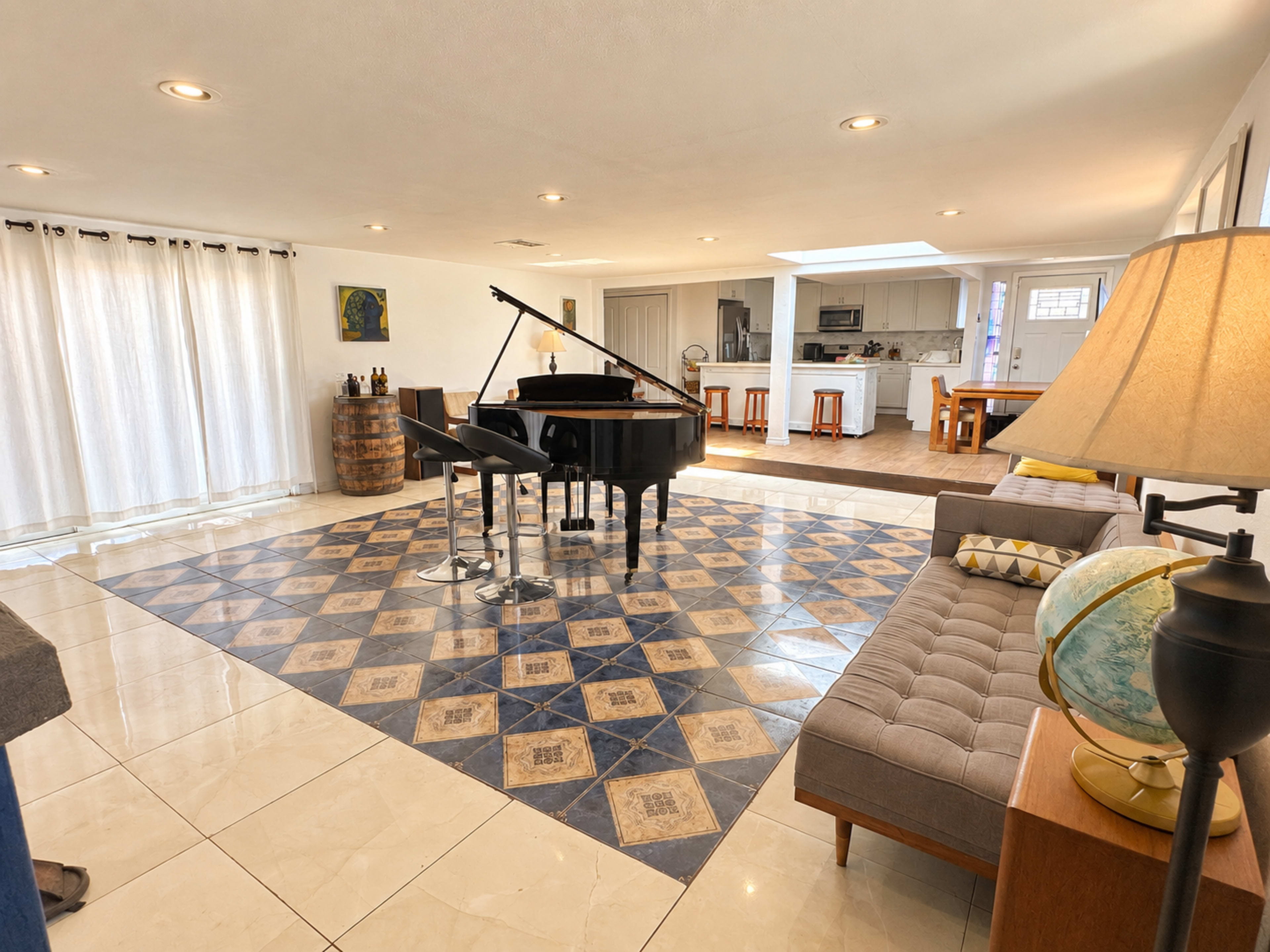 The image shows a spacious living area featuring a grand piano on an intricately tiled floor, surrounded by a sectional sofa and a wooden coffee table, with a kitchen area visible in the background.