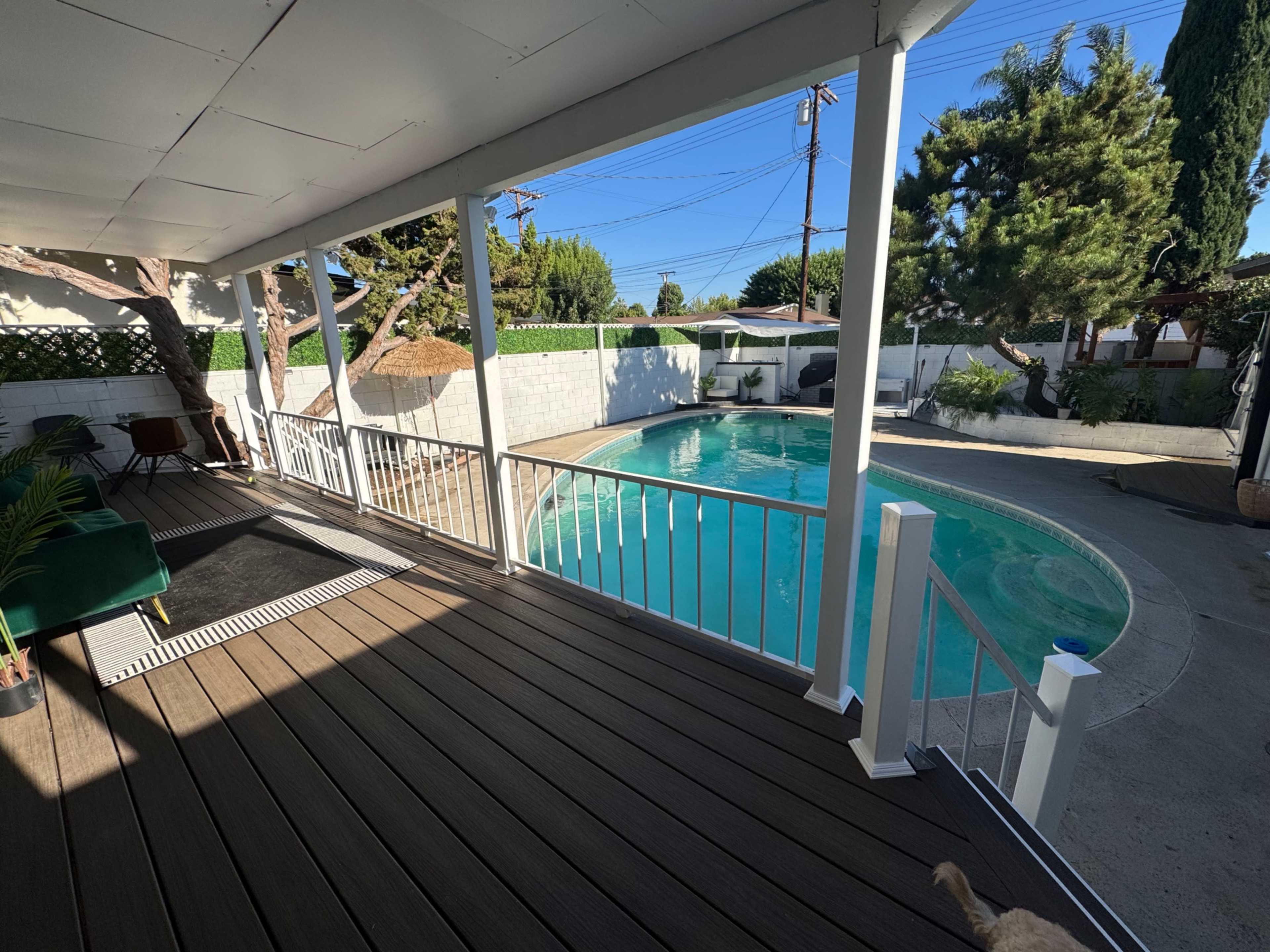 The image shows a backyard with a swimming pool surrounded by concrete and greenery, viewed from a covered porch.