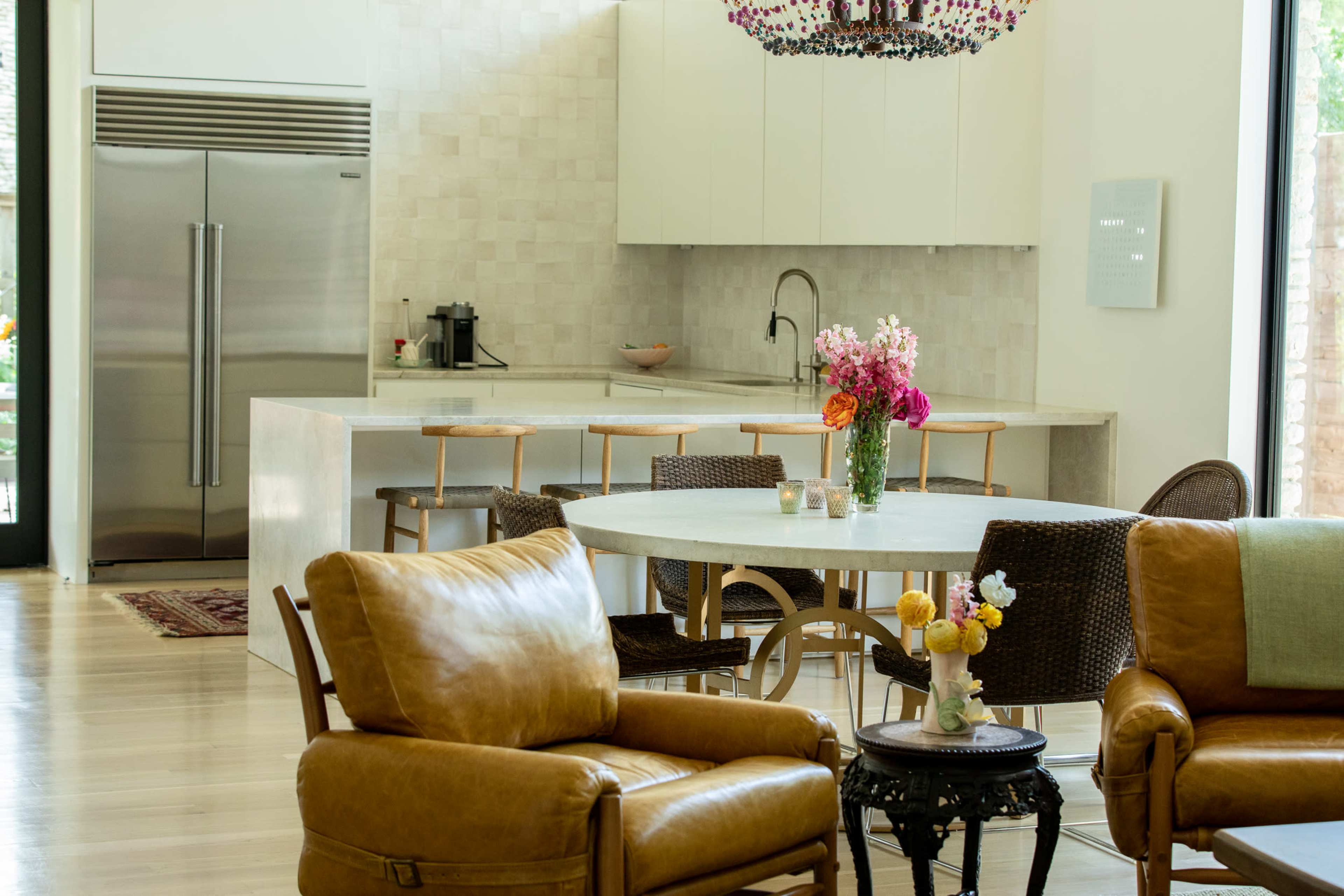 A bright, modern kitchen with a large island and dining table, surrounded by stylish seating, features a bouquet of flowers on the table.