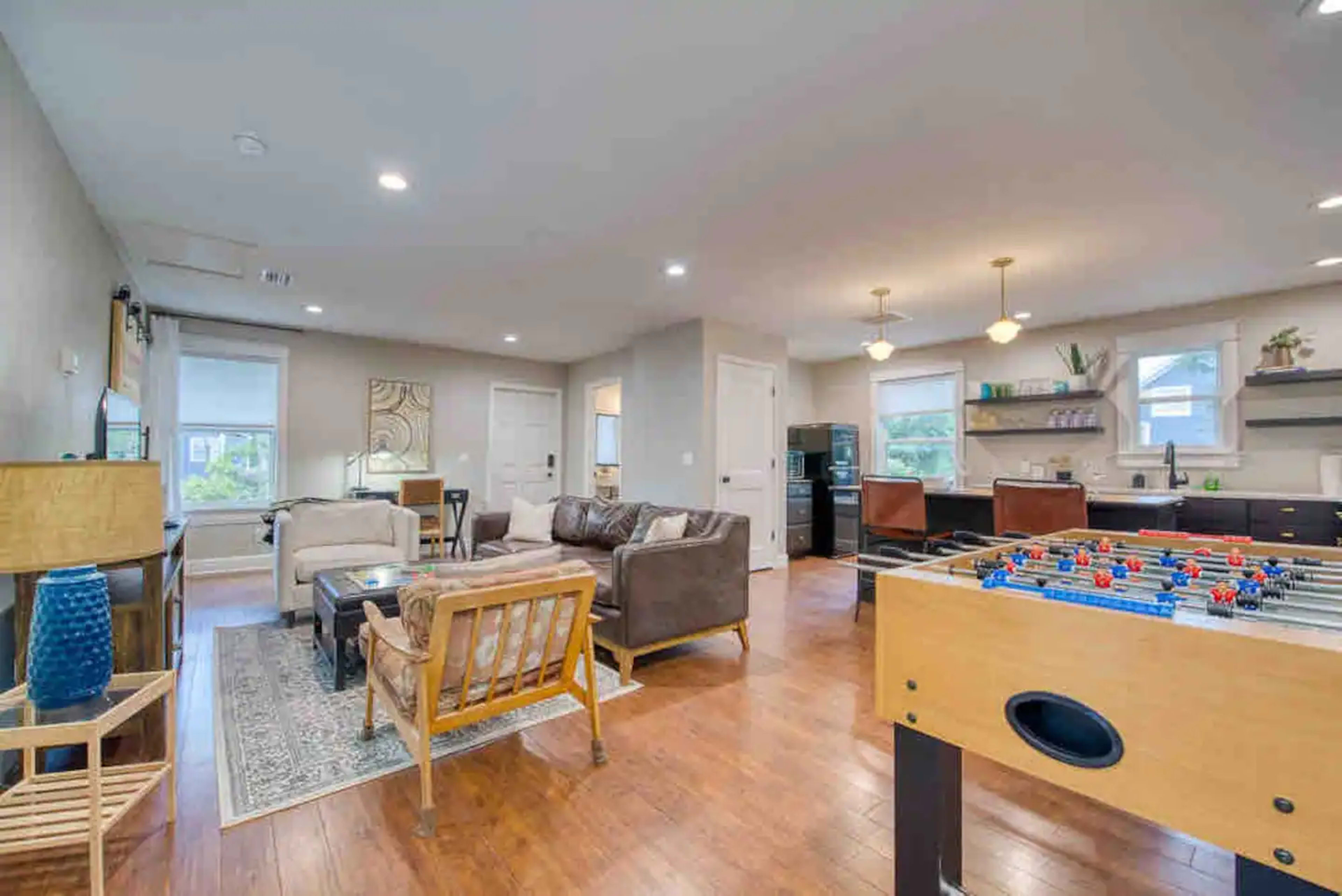 A modern living space featuring a foosball table, a seating area with two sofas, a coffee table, and a kitchen in the background.