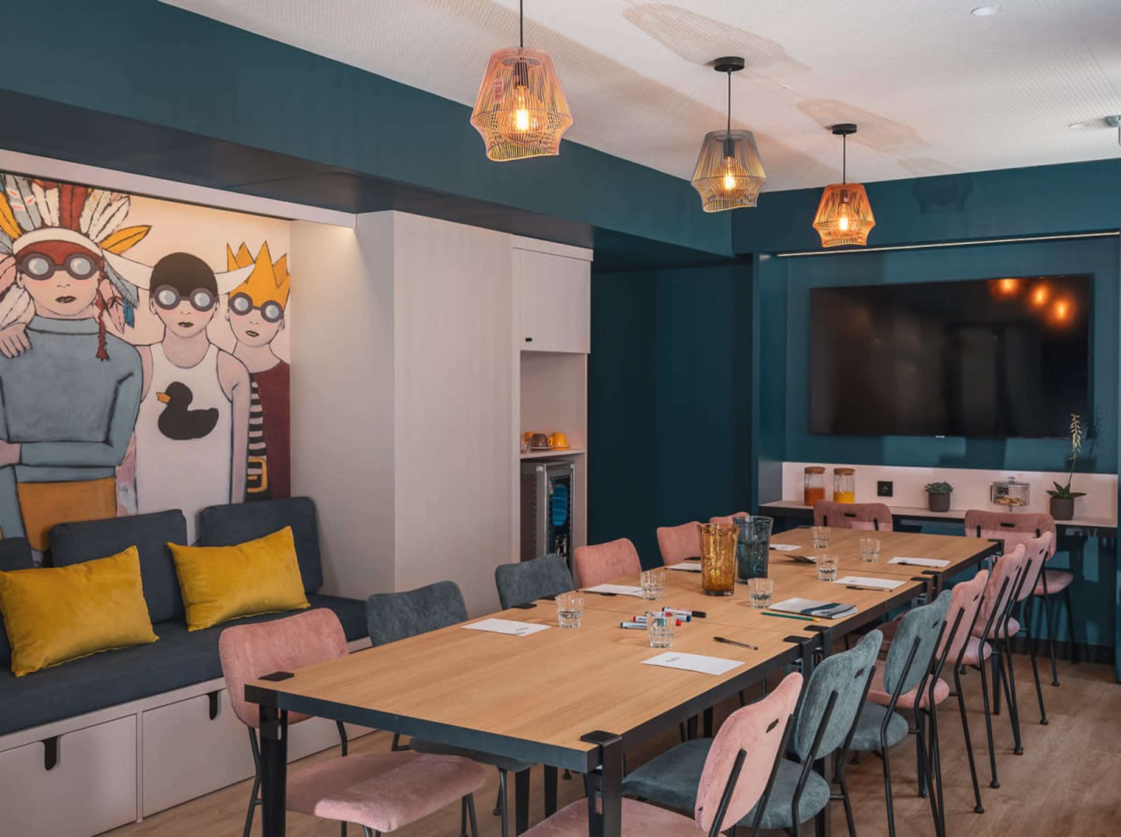 The image shows a modern meeting room featuring a long wooden table surrounded by variously colored chairs, with a decorative wall mural and a television on the opposite wall.