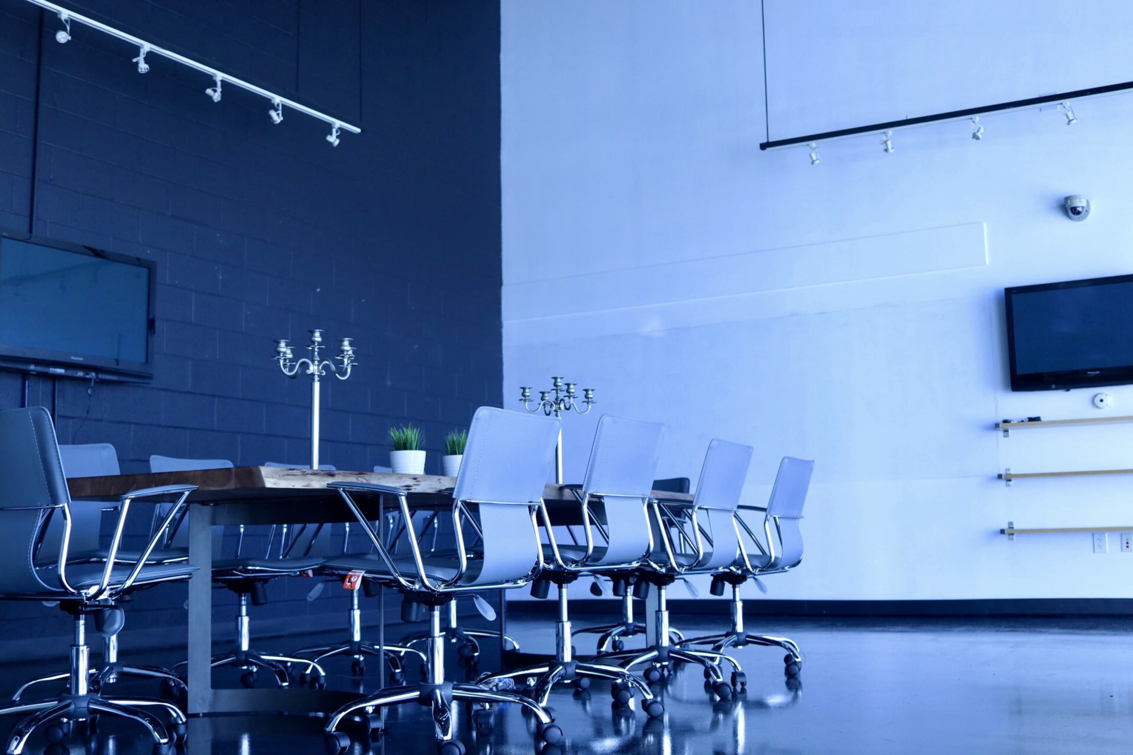 A modern conference room features a large table surrounded by chrome chairs, with a minimalist design and two televisions mounted on the walls.