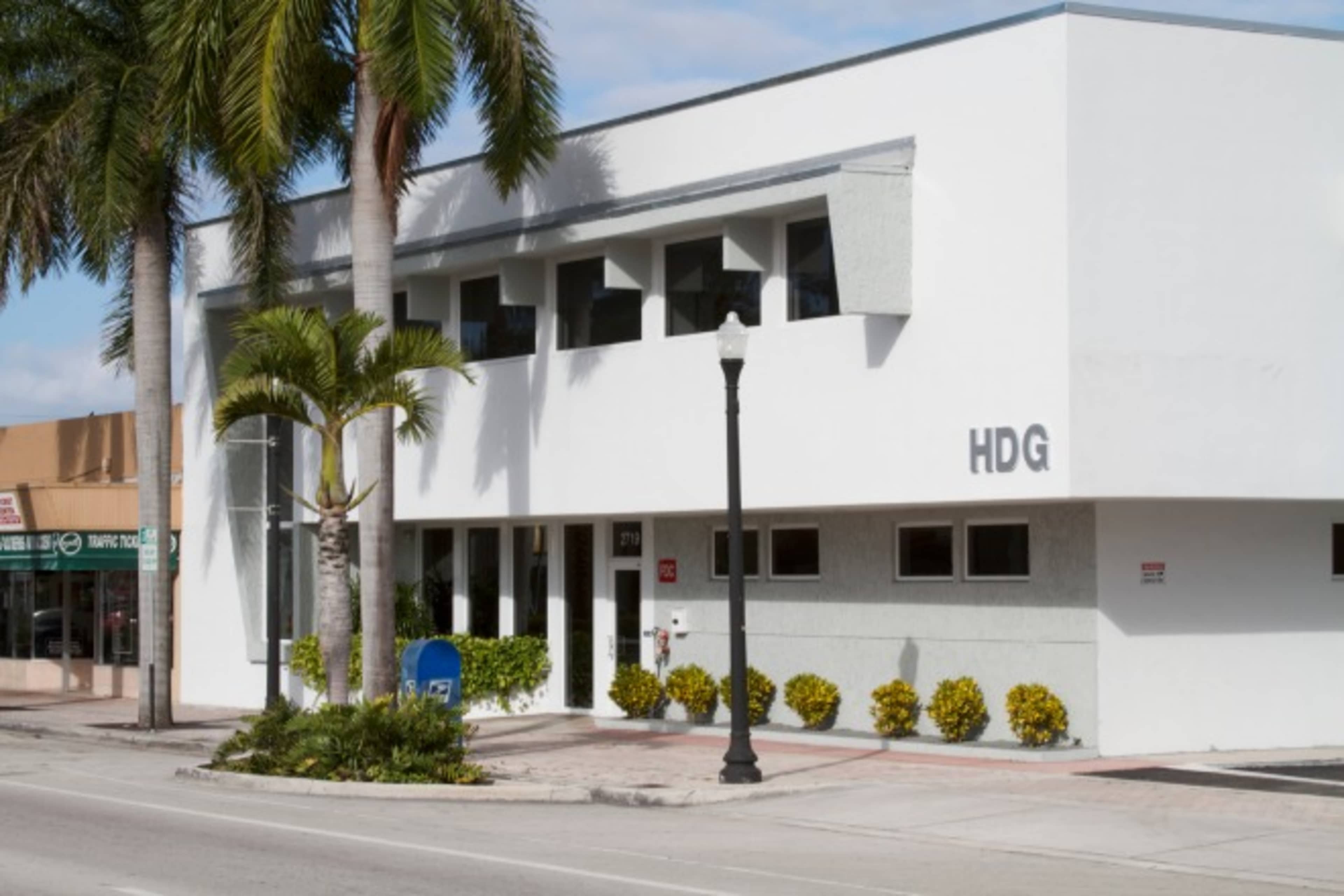 The image shows a modern white building with large windows, palm trees in front, and a sign that reads "HDG."