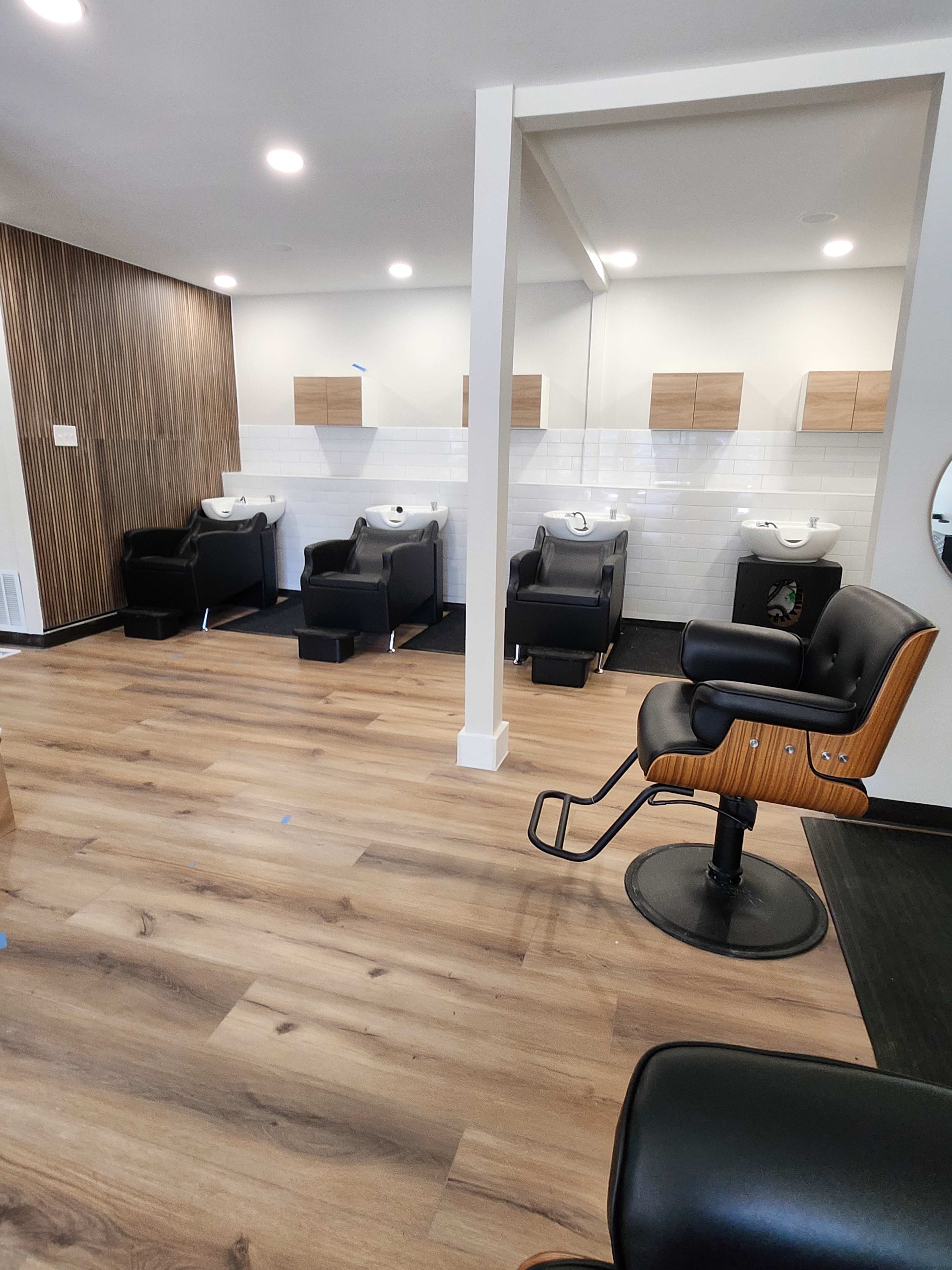 The image shows a modern salon interior with several black styling chairs and shampoo stations against a wooden and white tiled wall.