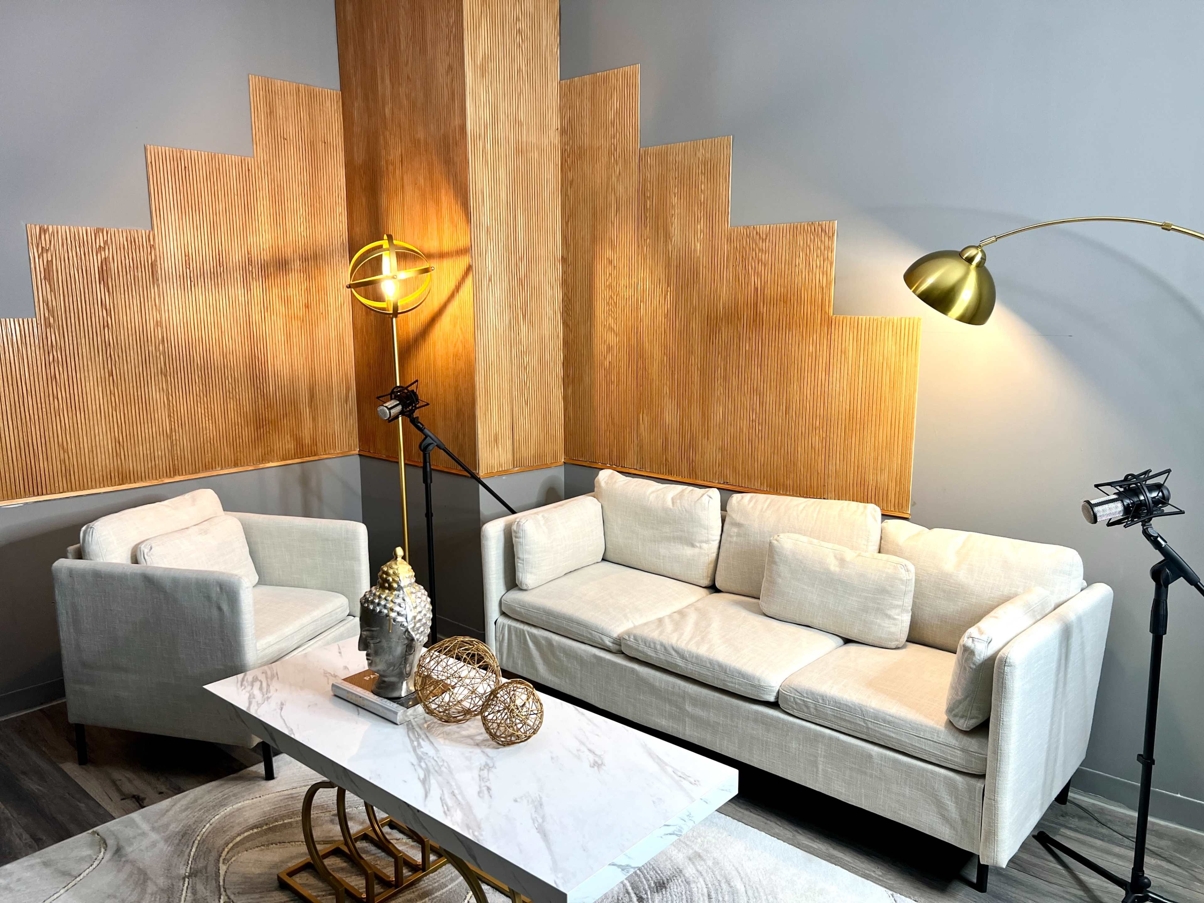 A modern seating area with a light-colored sofa and armchair, a marble table, and decorative elements, illuminated by a floor lamp and wall-mounted lights.