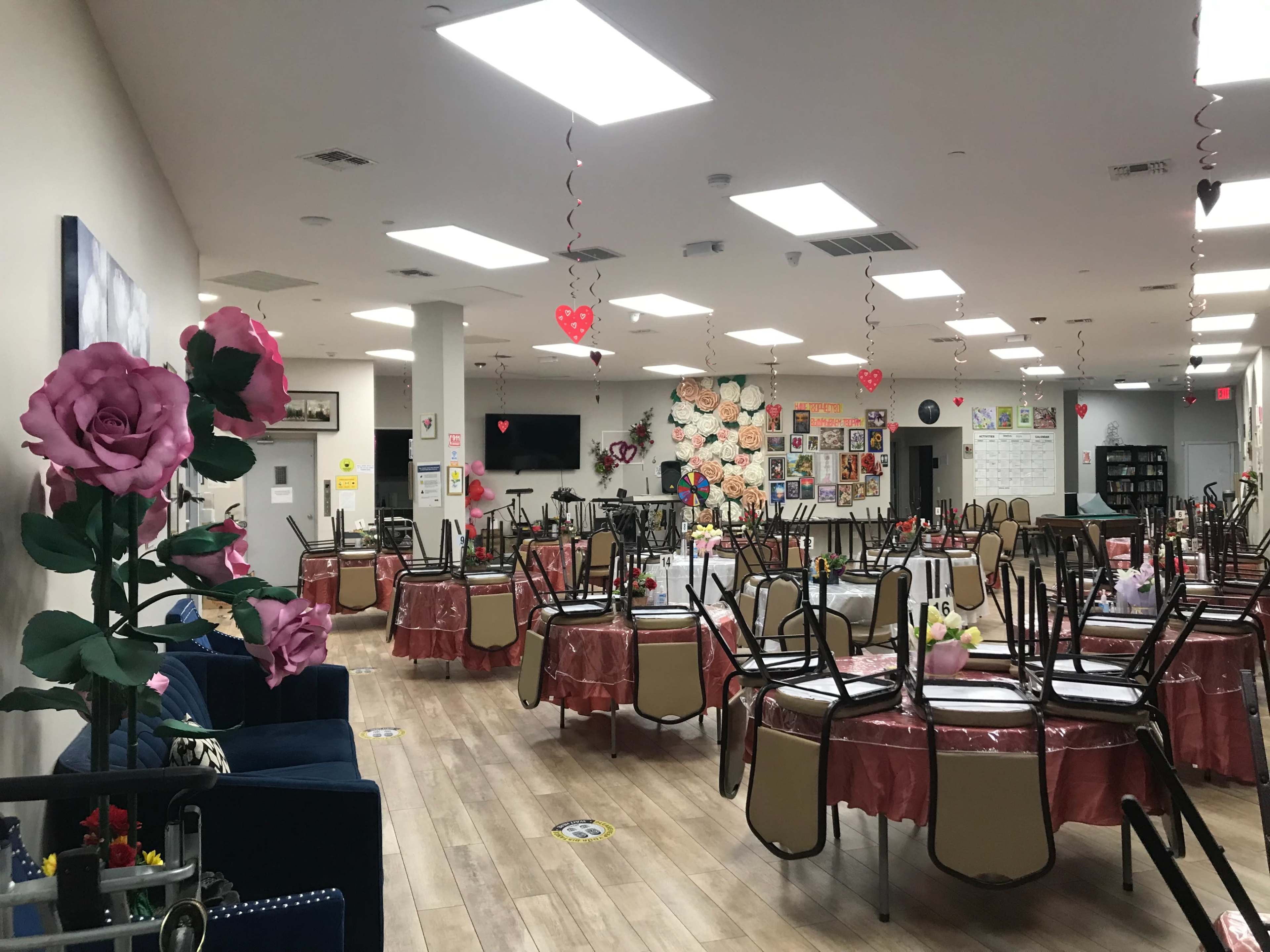 A large, decorated dining area with tables set for a gathering, featuring floral centerpieces and heart-shaped decorations hanging from the ceiling.