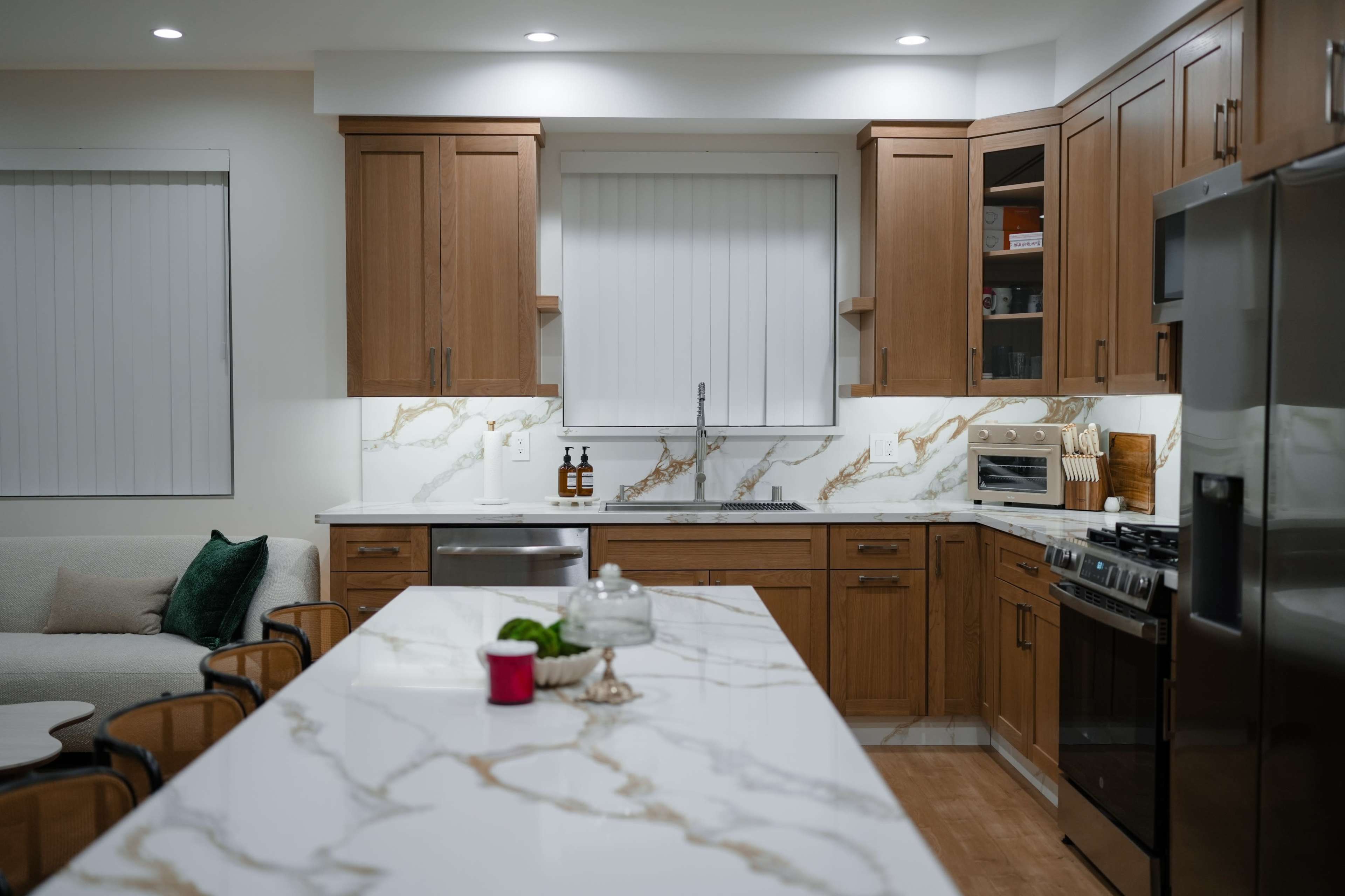 A modern kitchen features wooden cabinets, a marble countertop, and stainless steel appliances, along with a cozy seating area in the background.