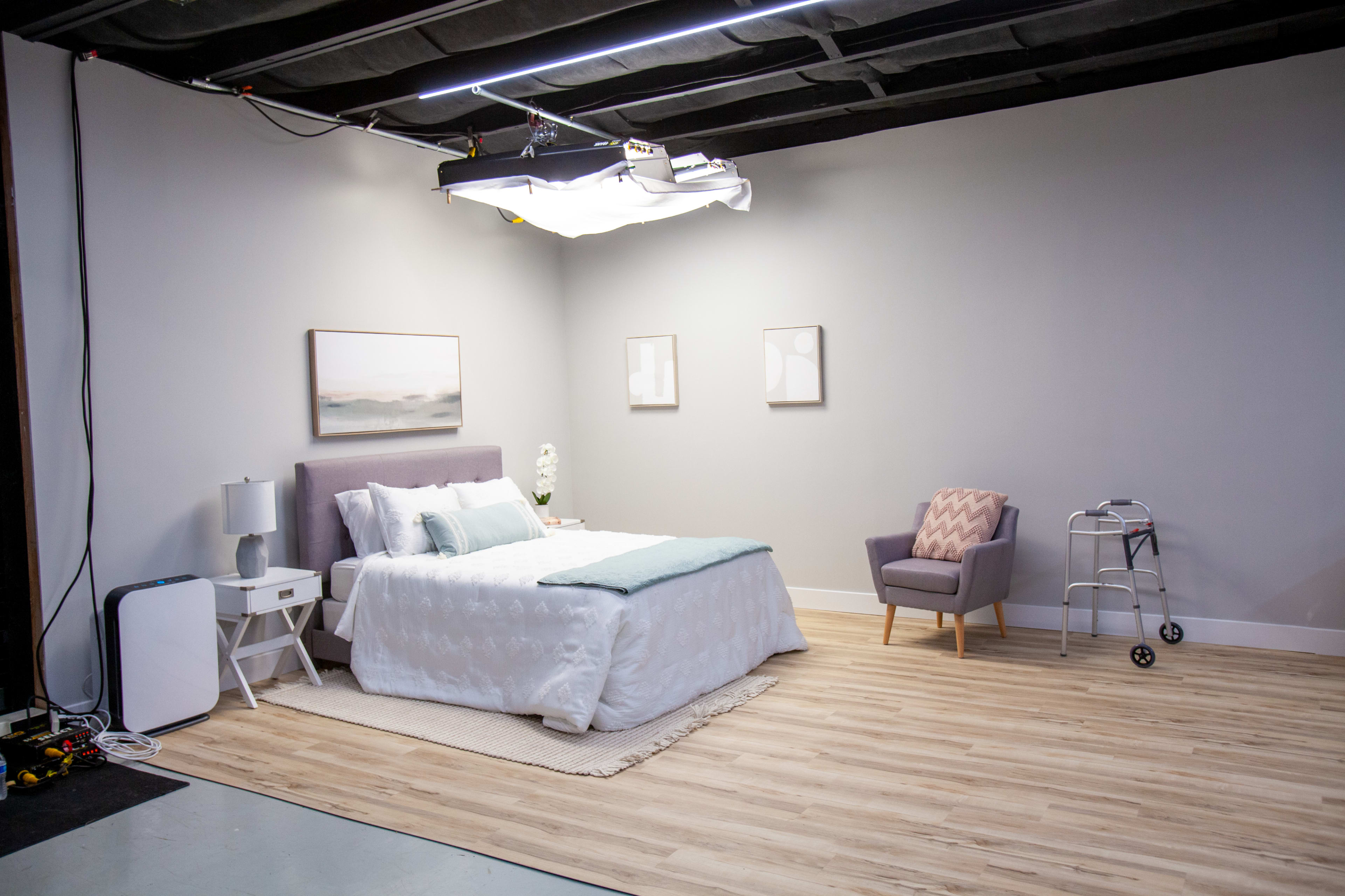 A minimalistic bedroom setup featuring a bed with a light blue comforter, a nightstand, a chair, and a walker in a well-lit space.