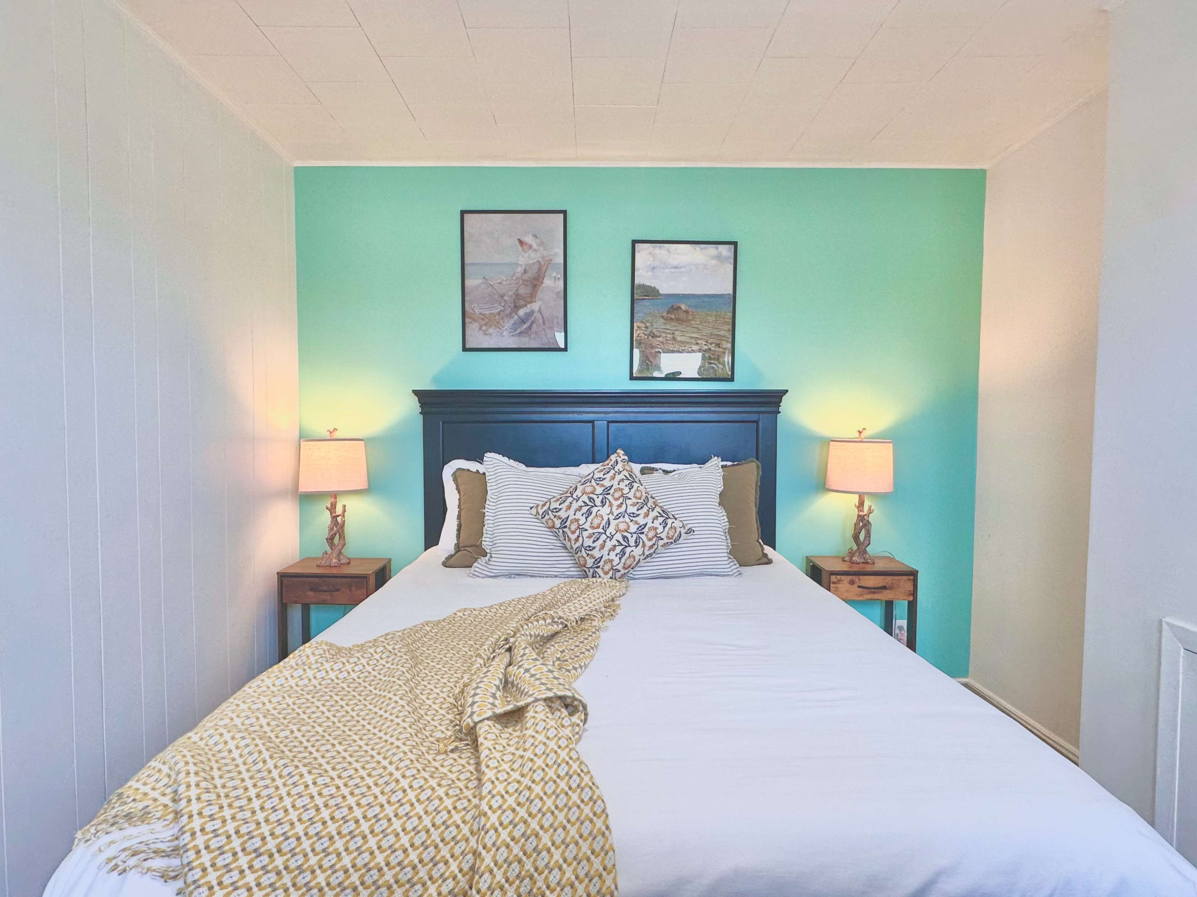 The image shows a neatly arranged bedroom with a bed, two bedside tables with lamps, and framed artwork on a teal wall.