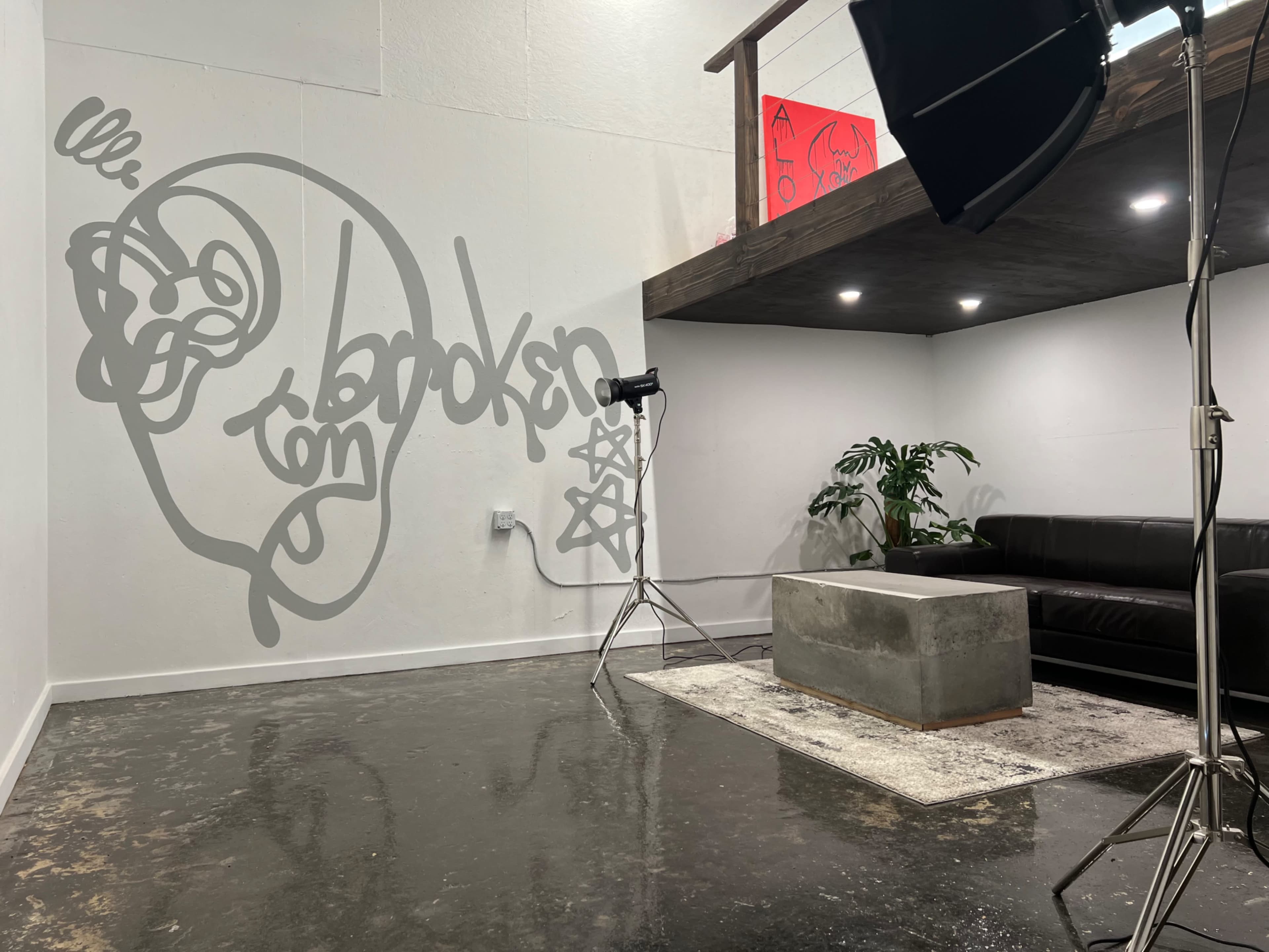 A modern interior with a black couch, a concrete coffee table, a photography light, and a large mural of a stylized face on the wall.