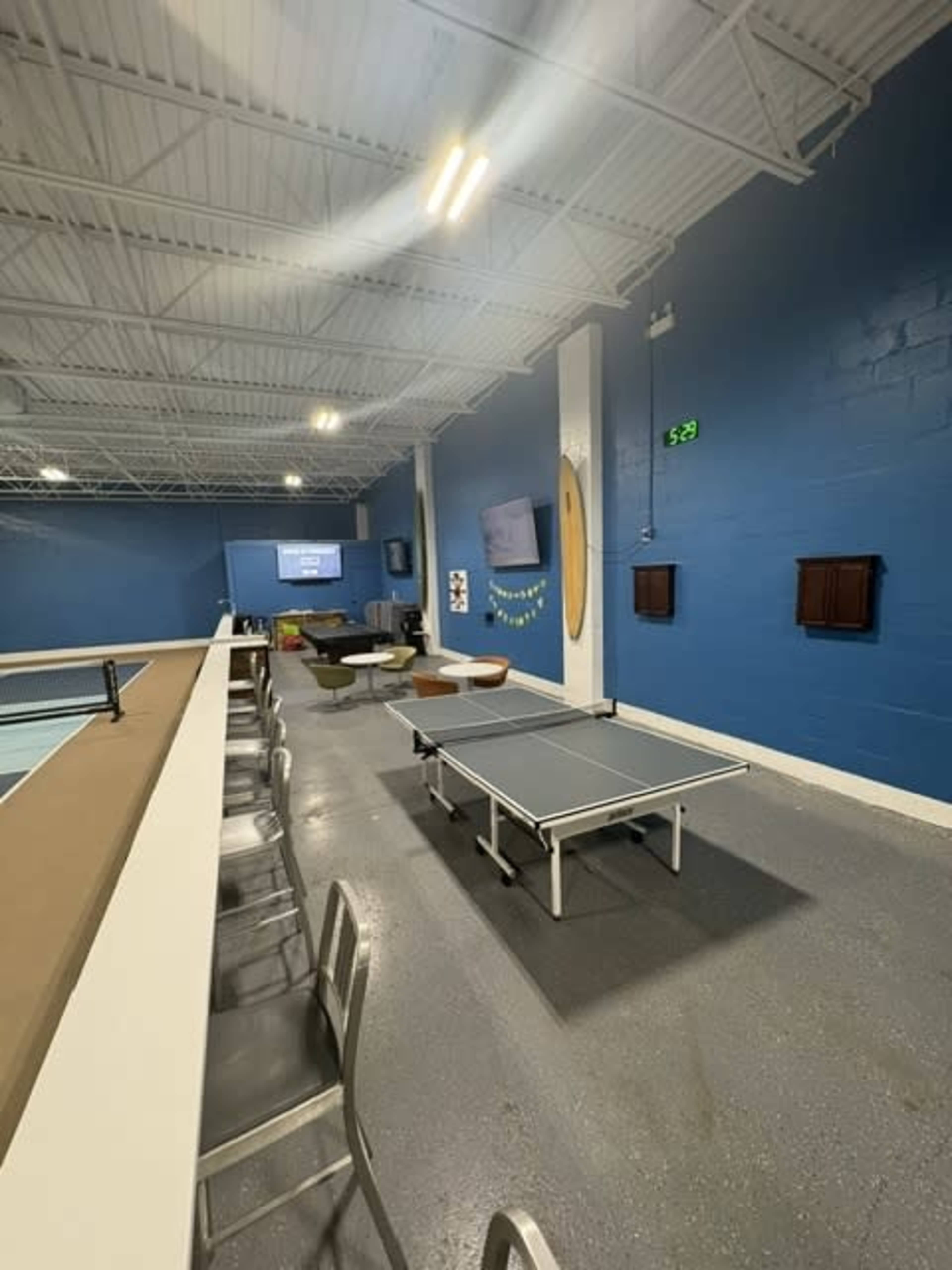 The image shows a recreational room featuring a ping pong table, a bar with stools, seating areas, and blue walls.