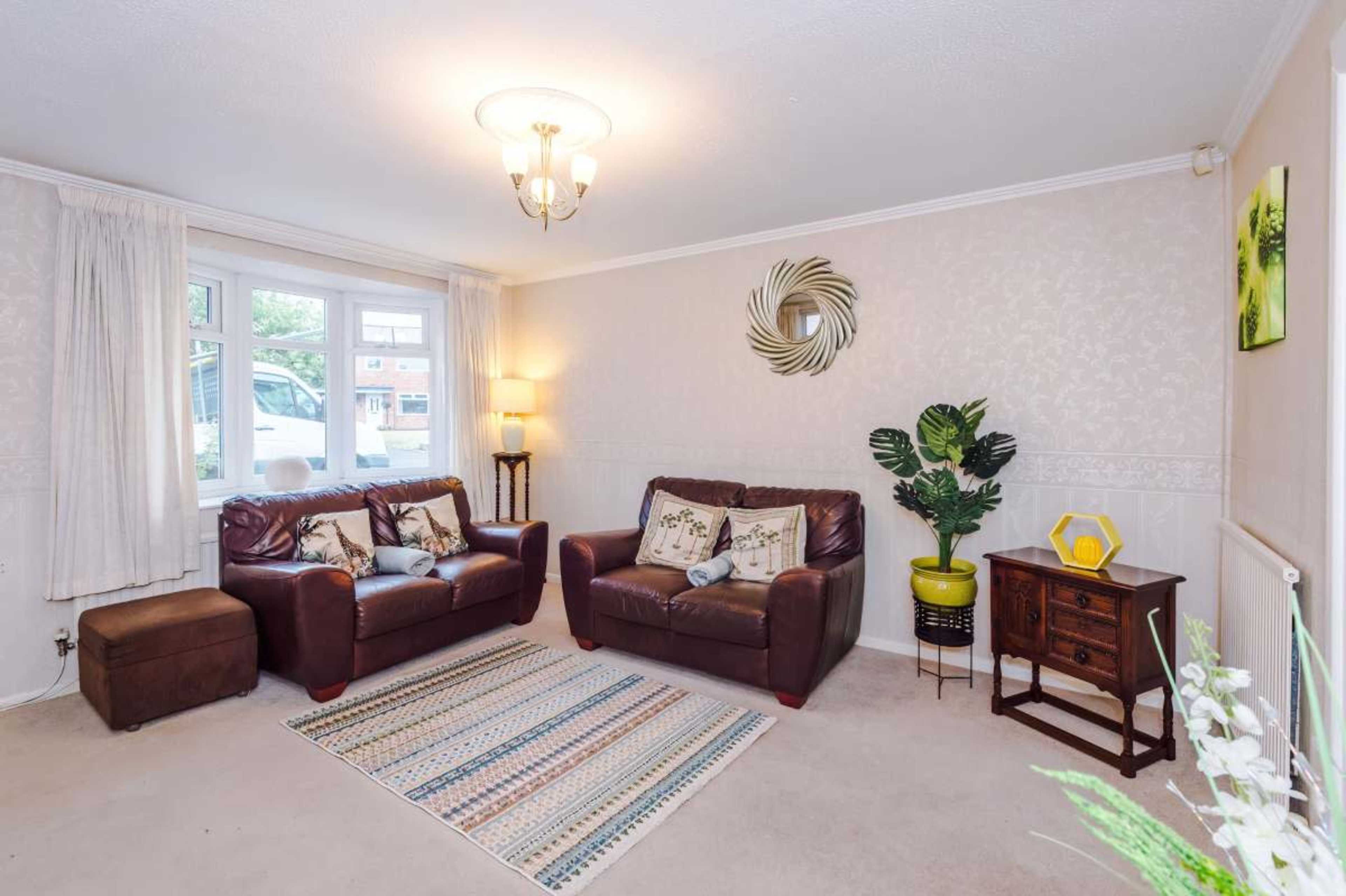 The image shows a cozy living room with two brown leather sofas, a small wooden side table, a plant in a yellow pot, and a patterned rug on the carpeted floor.