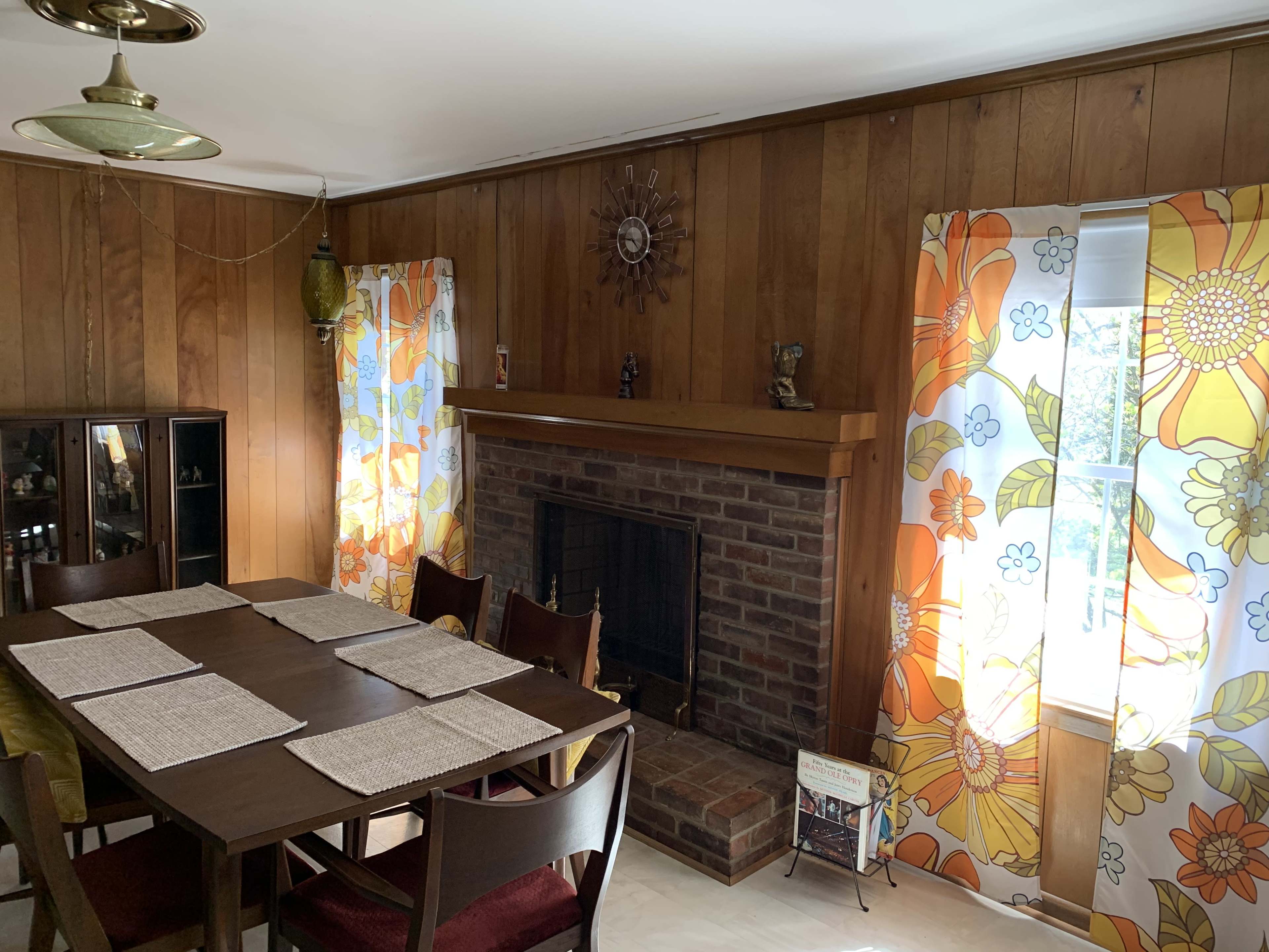 The image depicts a dining area with a wooden table, chairs, a brick fireplace, and floral-patterned curtains on the windows.
