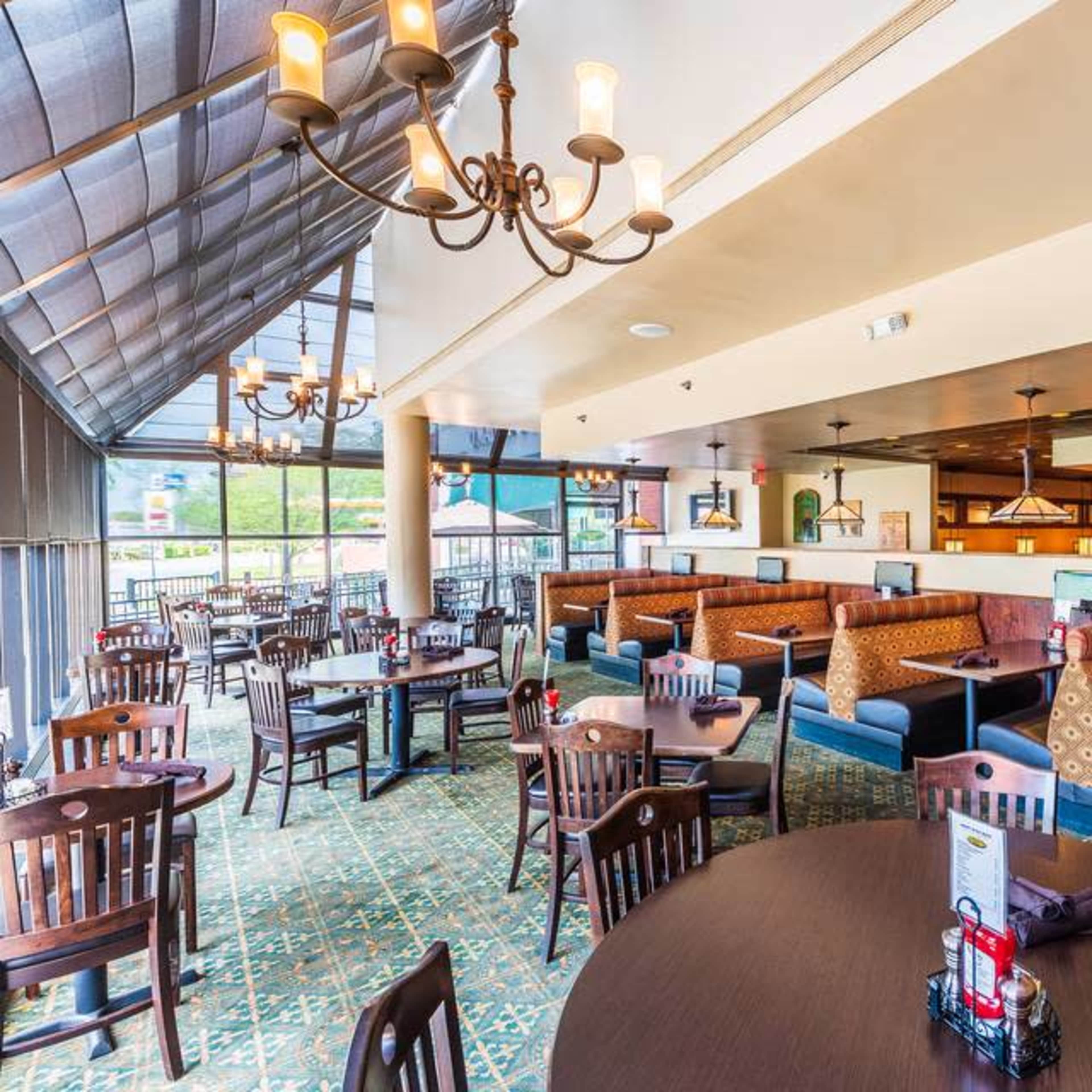 A restaurant interior with wooden tables, chairs, and booths arranged beneath a high ceiling with chandeliers and large windows allowing natural light.