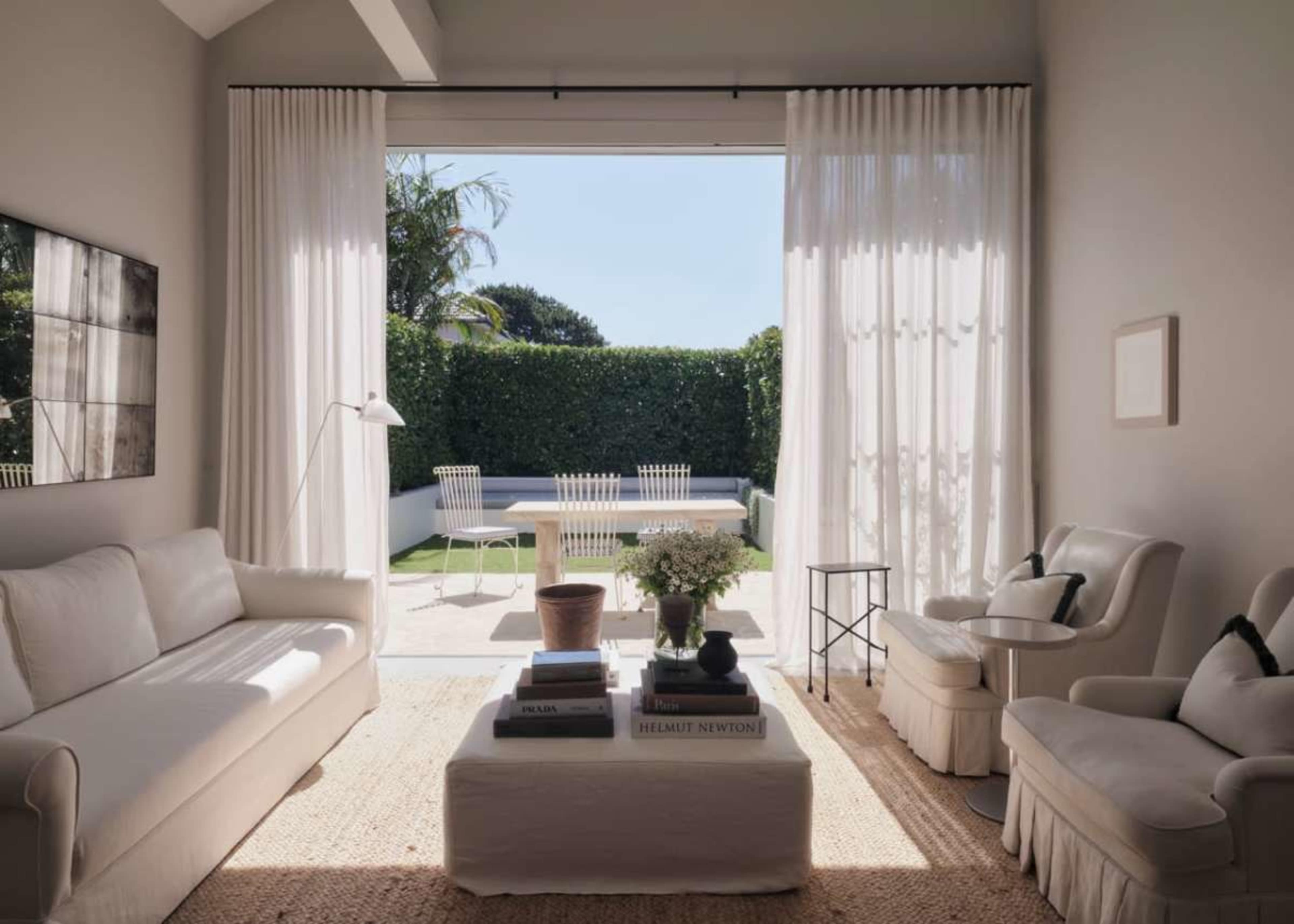 A bright living room features a white sofa, two armchairs, and large windows that open to a garden with a swimming pool.