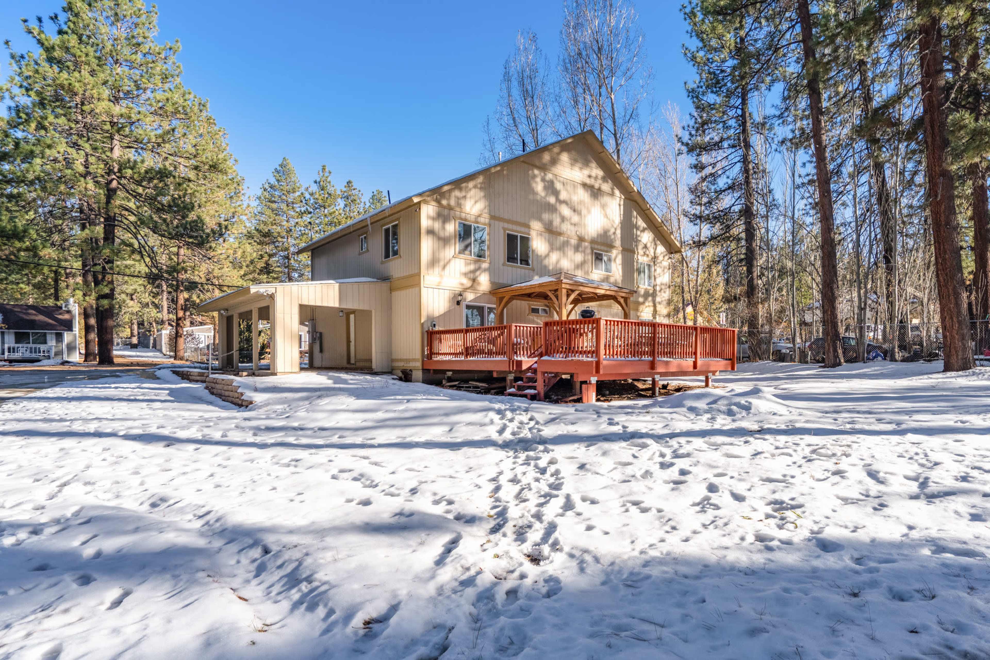 A two-story house with a red deck stands among snow-covered ground and tall pine trees.