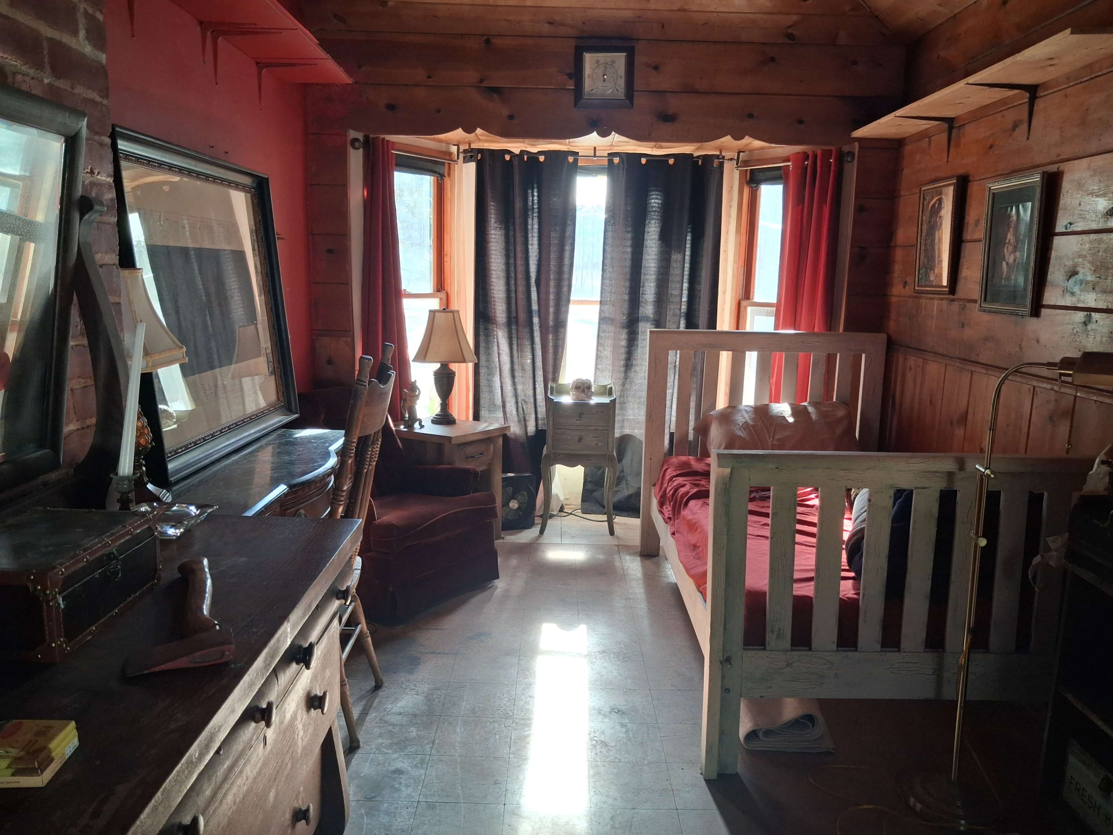 The image shows a cozy wooden bedroom with a bed, a table with a lamp, and large windows dressed with dark curtains.