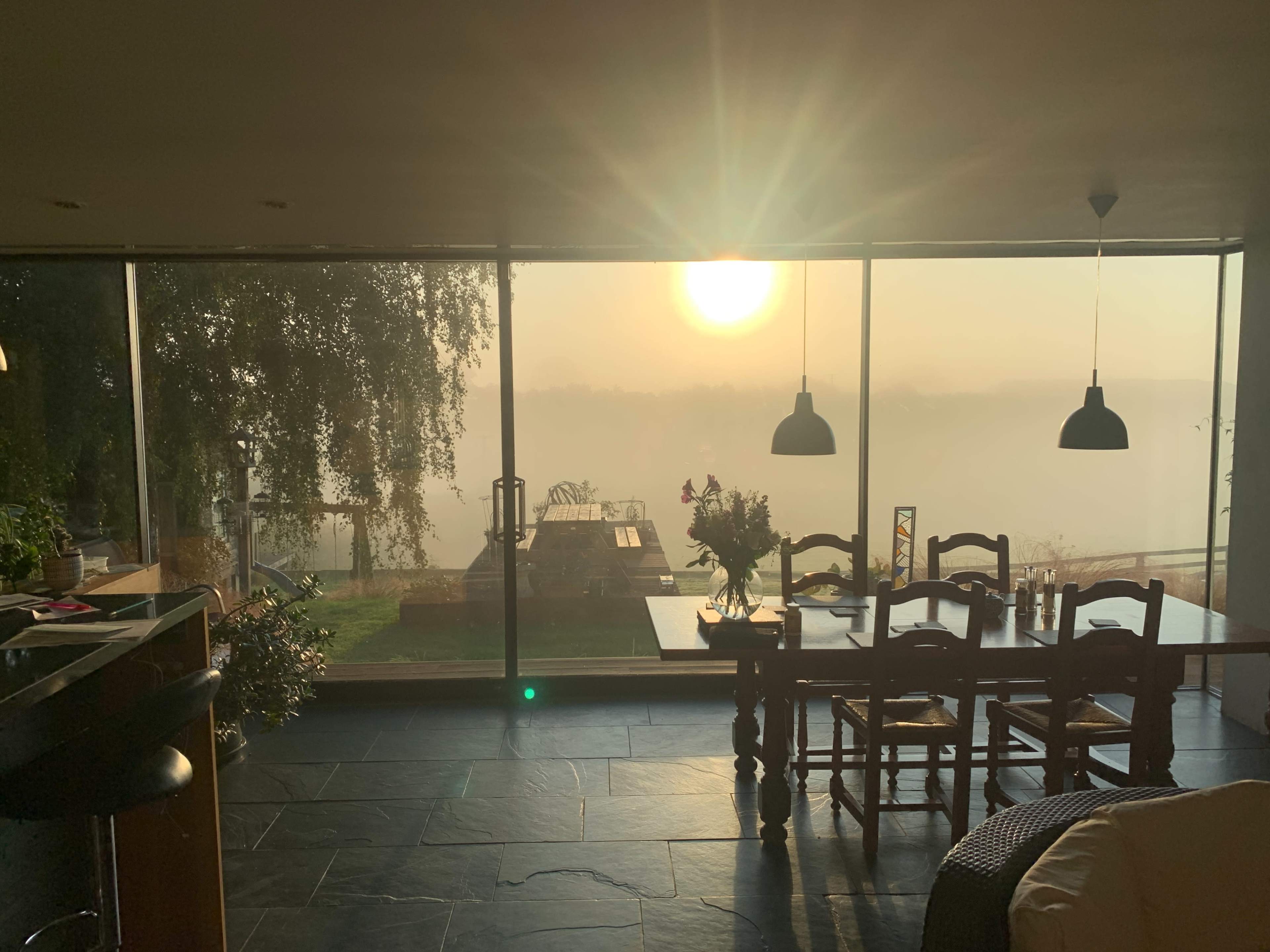 A sunlit dining area with a table and chairs overlooks a misty outdoor scene through large glass windows.