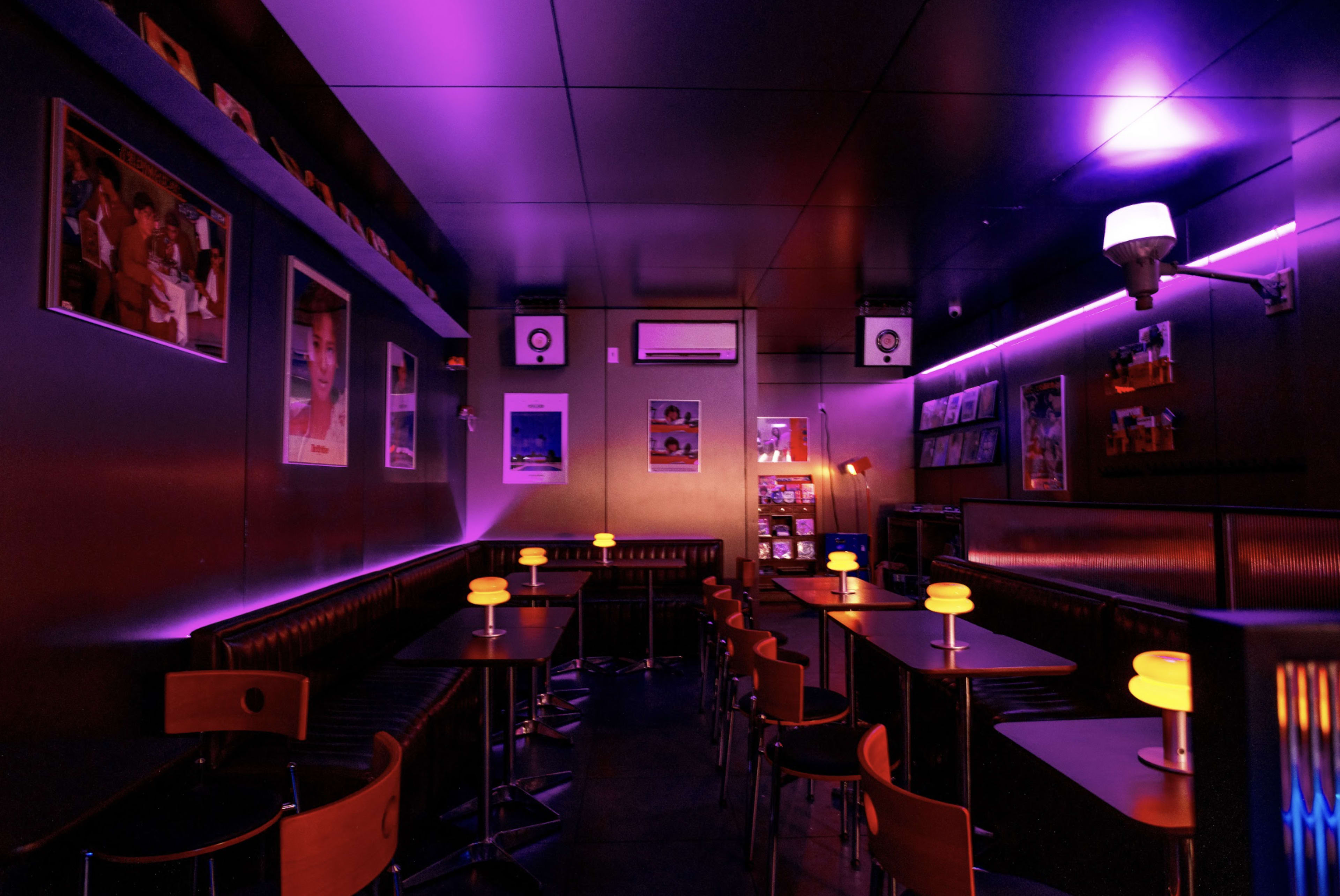 The image depicts a dimly lit bar interior with purple lighting, featuring black tables, chairs, and framed posters on the walls.