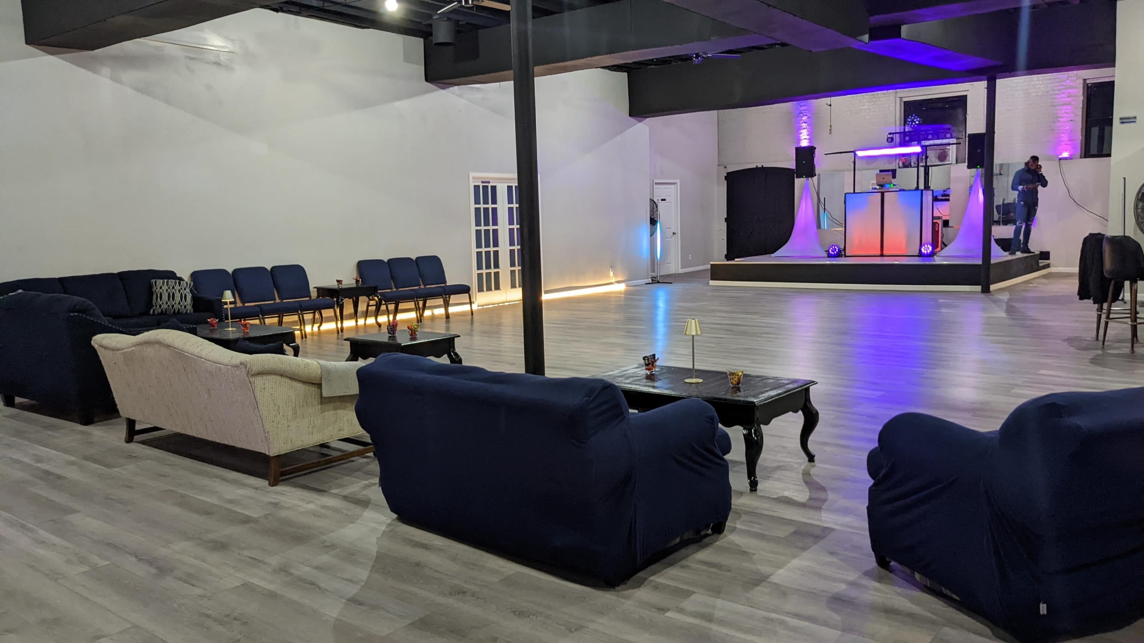 An empty event space with blue and purple lighting, featuring a stage, seating area with couches and chairs, and a bar setup in the background.