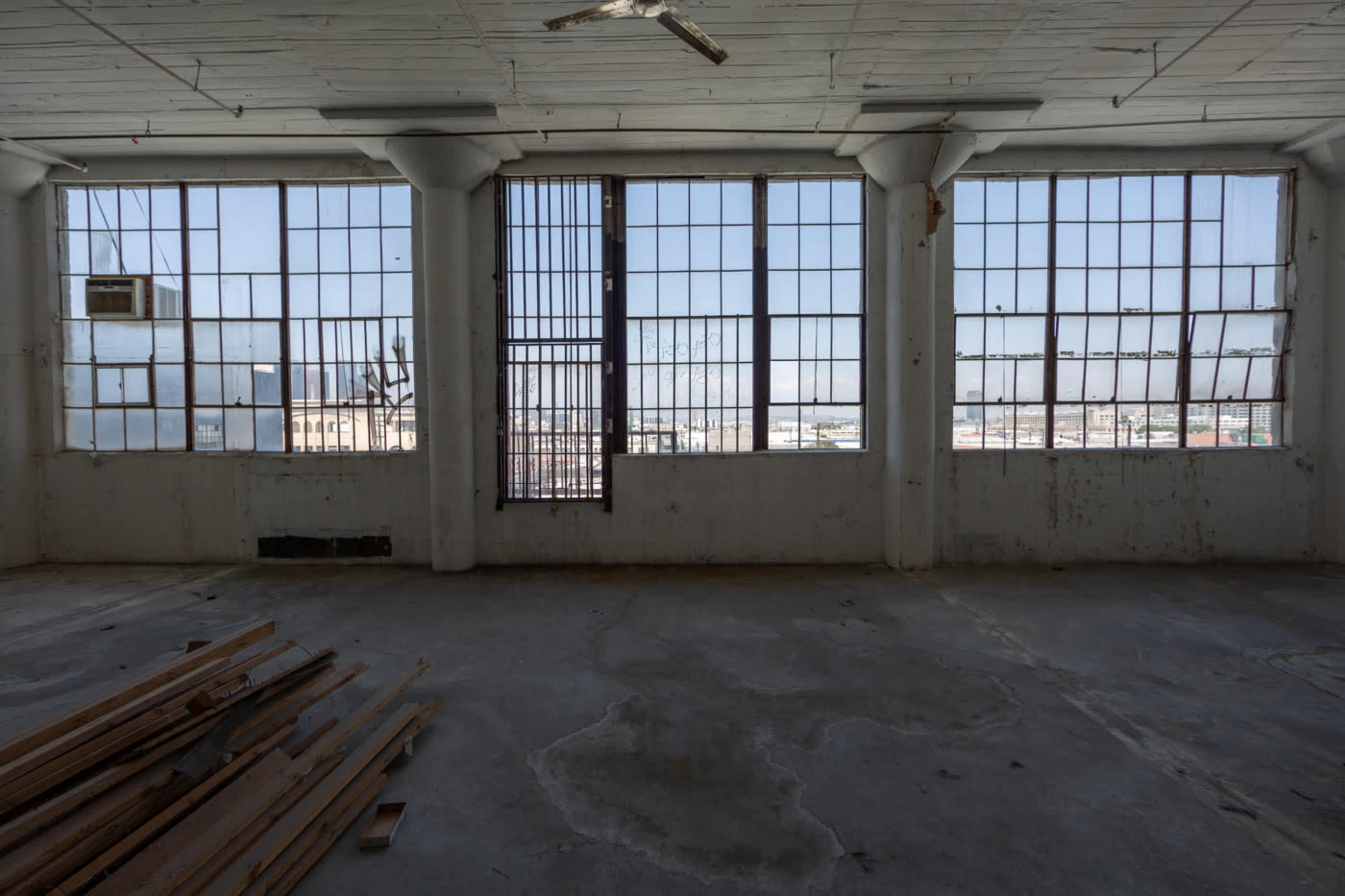 An empty industrial room with large, barred windows and piled wooden planks on the concrete floor.