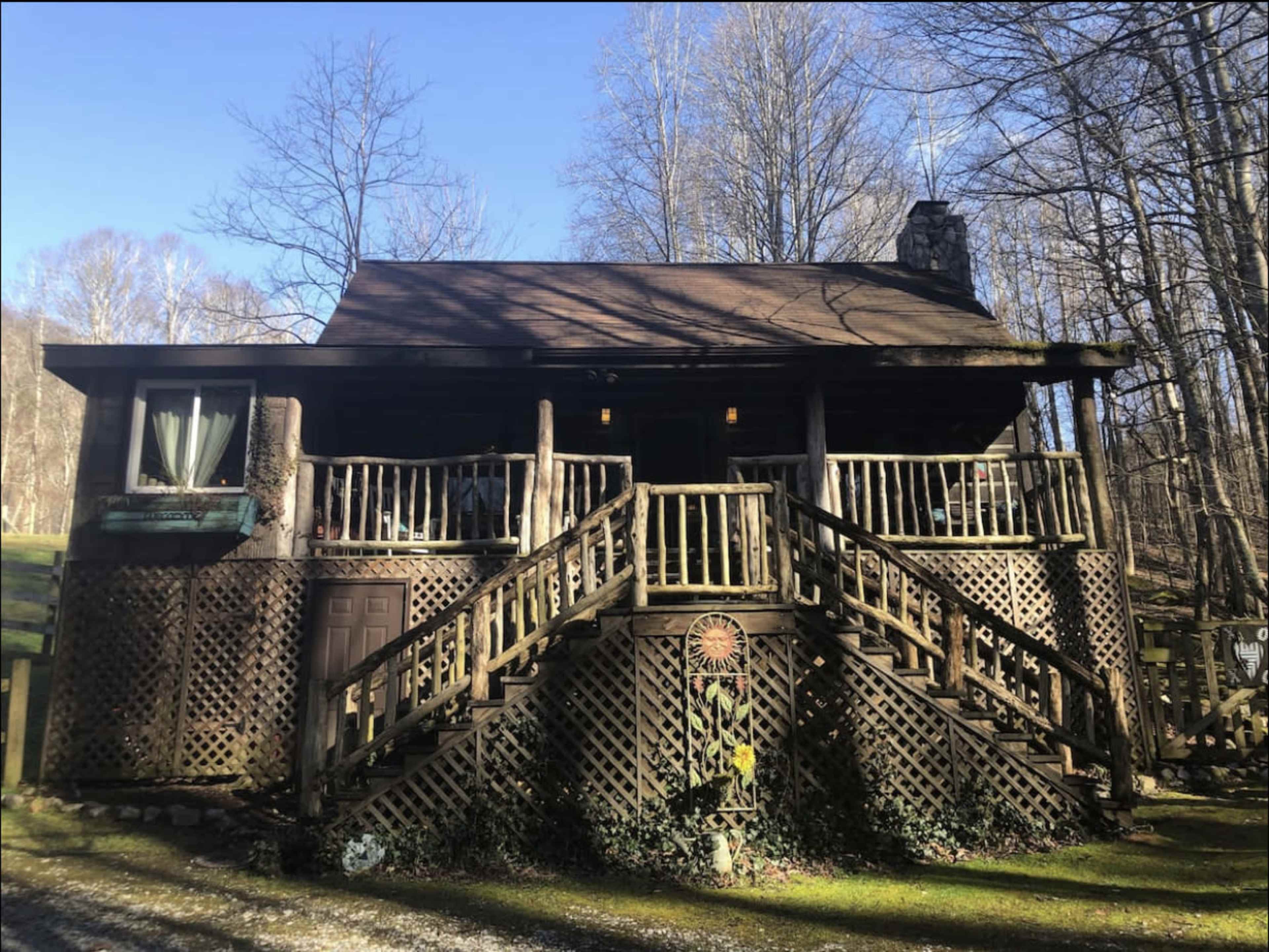 A rustic cabin with wooden lattice and two sets of stairs is surrounded by trees in a forested area.
