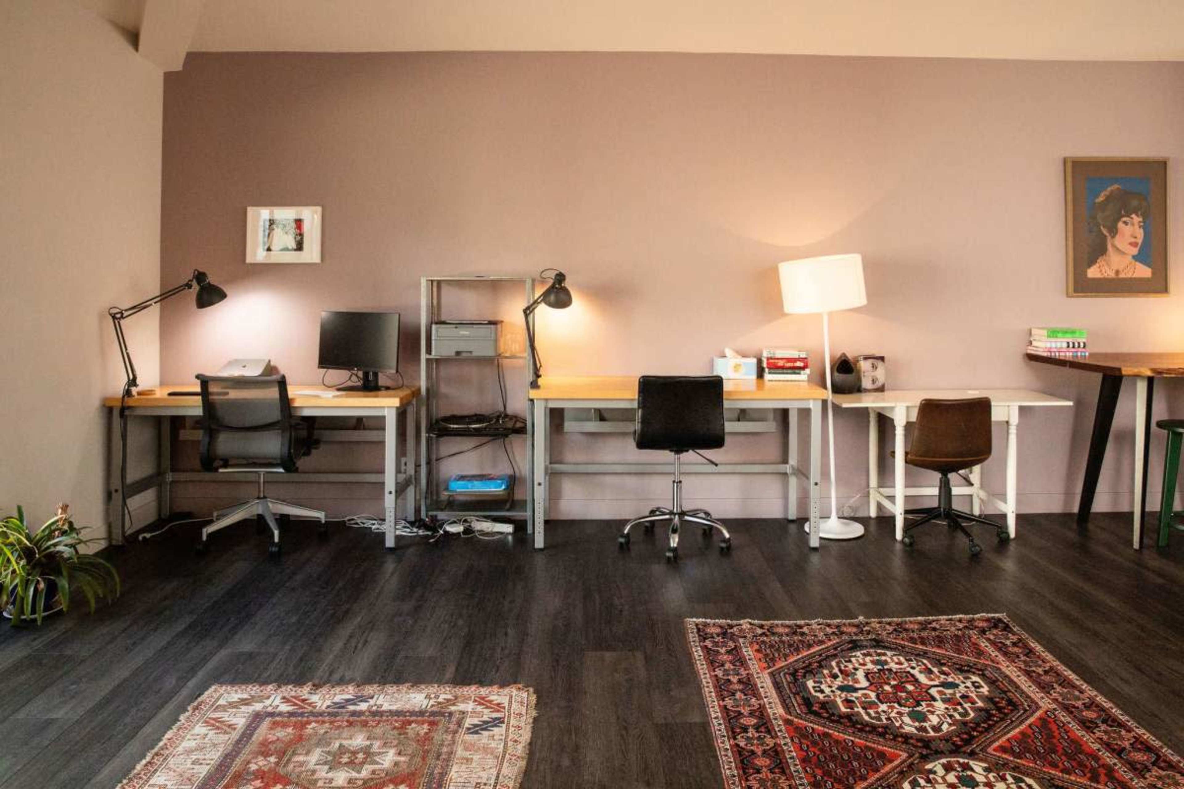 The image shows a spacious room with four desks, each with a chair, and a combination of floor and desk lamps, complemented by decorative rugs on a dark wooden floor.