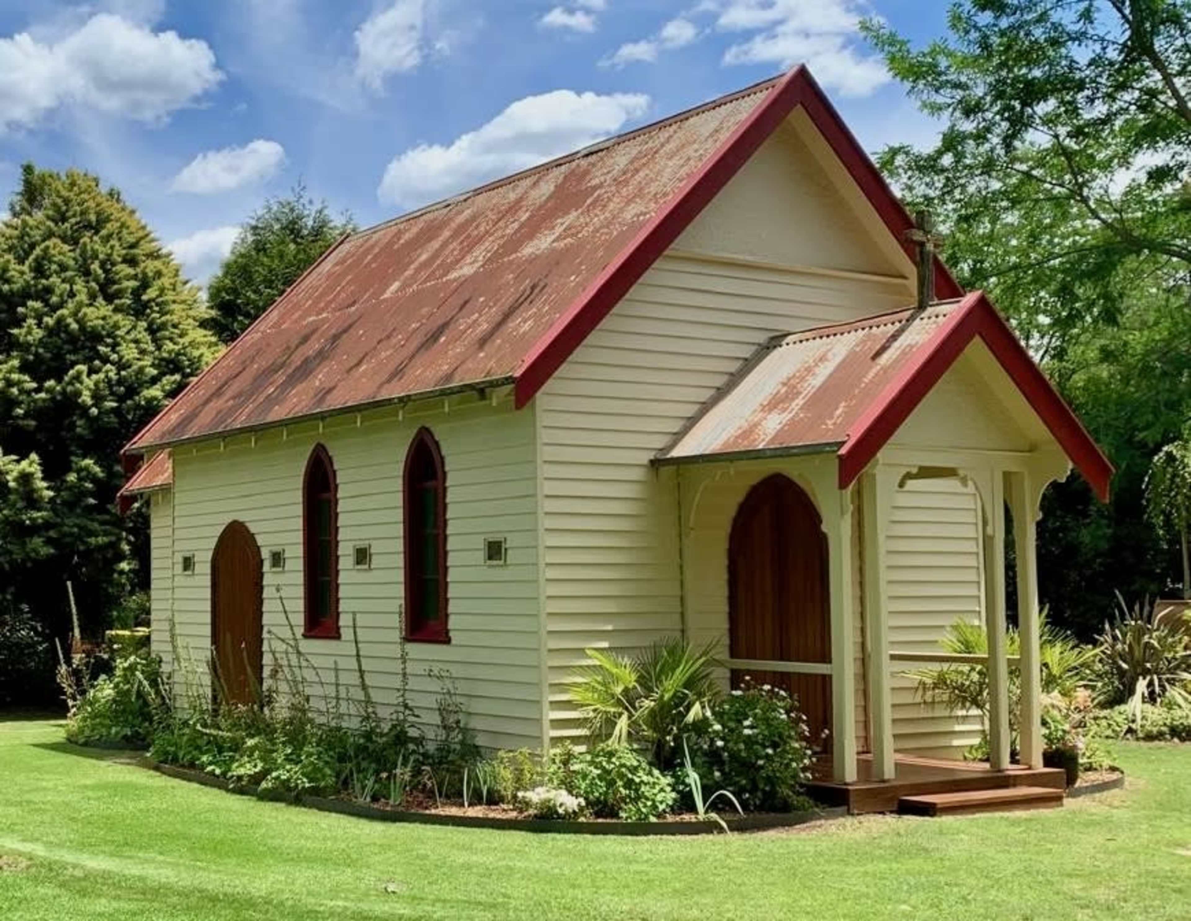 The image shows a small, rustic church building with a sloped roof and arched windows, surrounded by lush greenery in a well-kept garden.