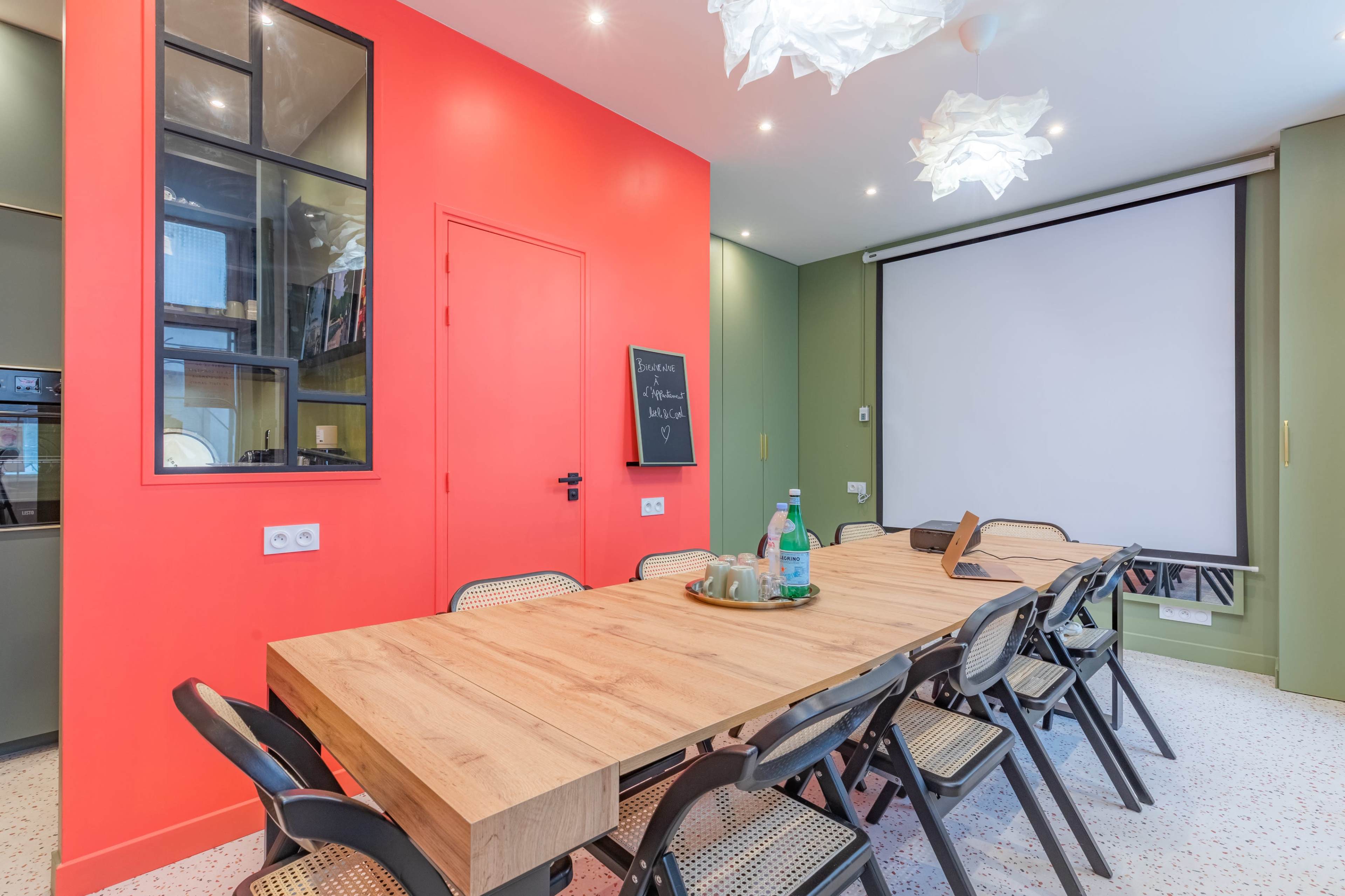 A modern meeting room features a long wooden table surrounded by black folding chairs, with red and green walls and a large screen for presentations.