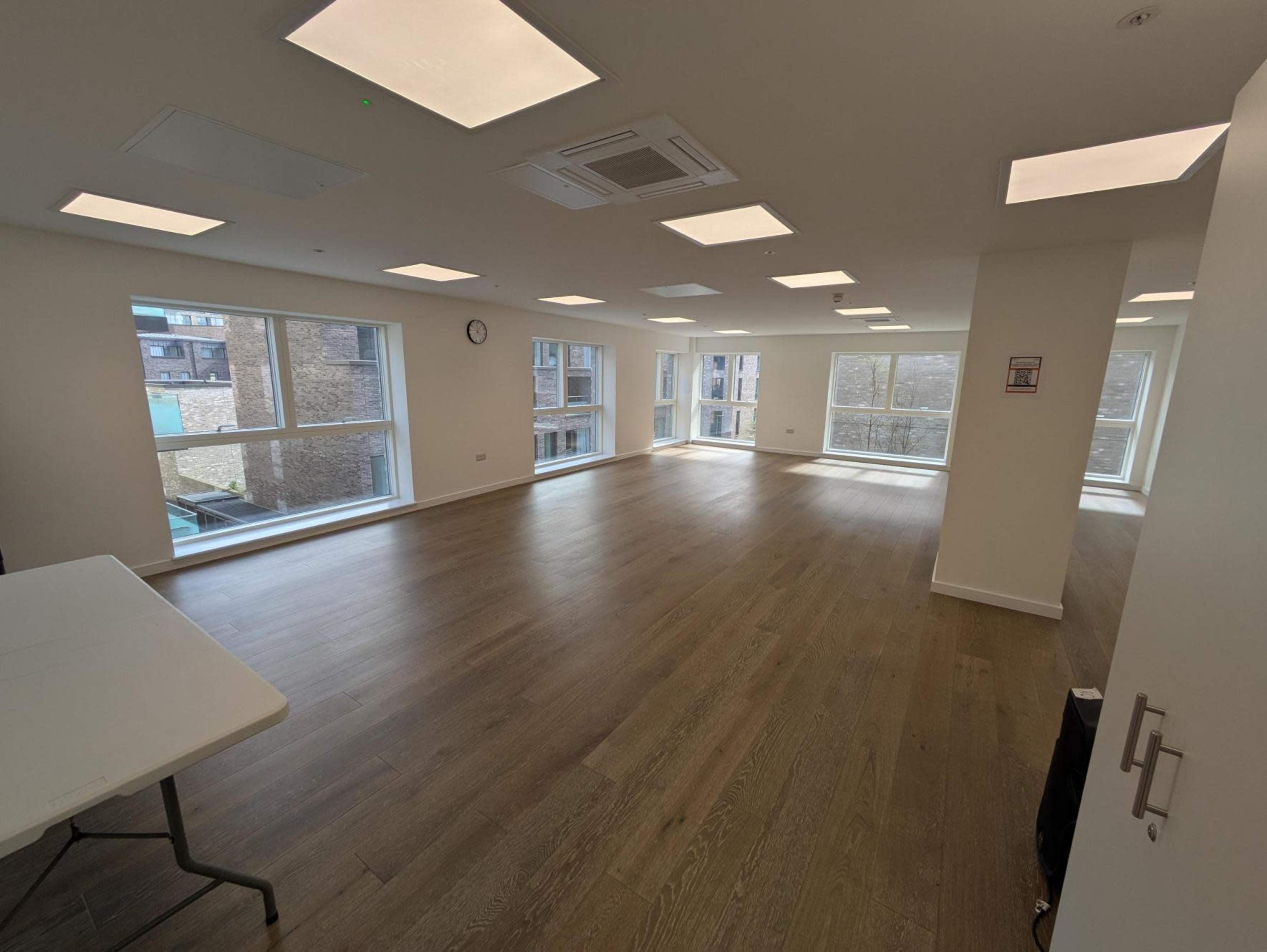 The image shows a spacious, empty office room with large windows and overhead ceiling lights.