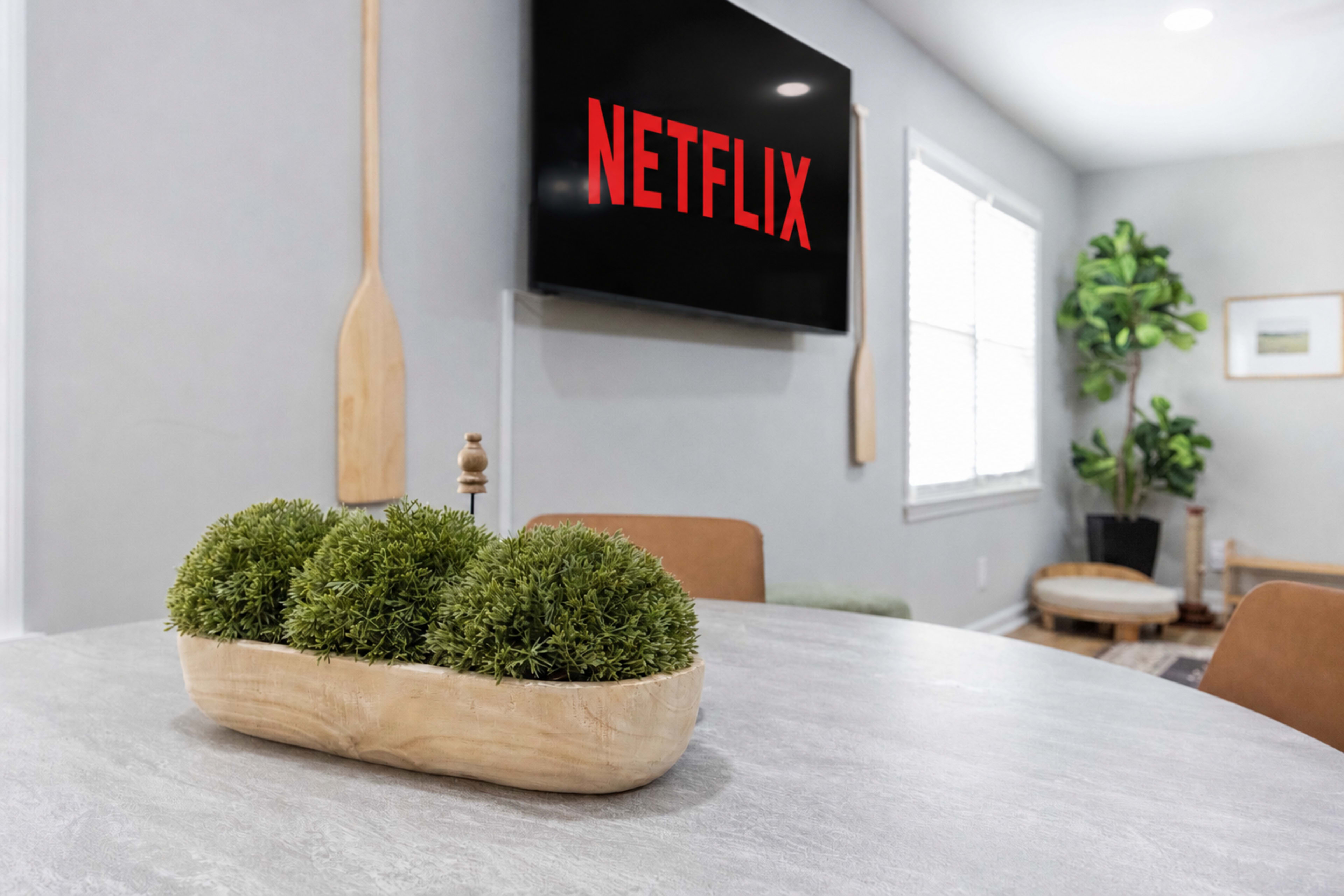 A modern living space features a circular table with a wooden bowl holding greenery, and a large television mounted on the wall displaying the Netflix logo.