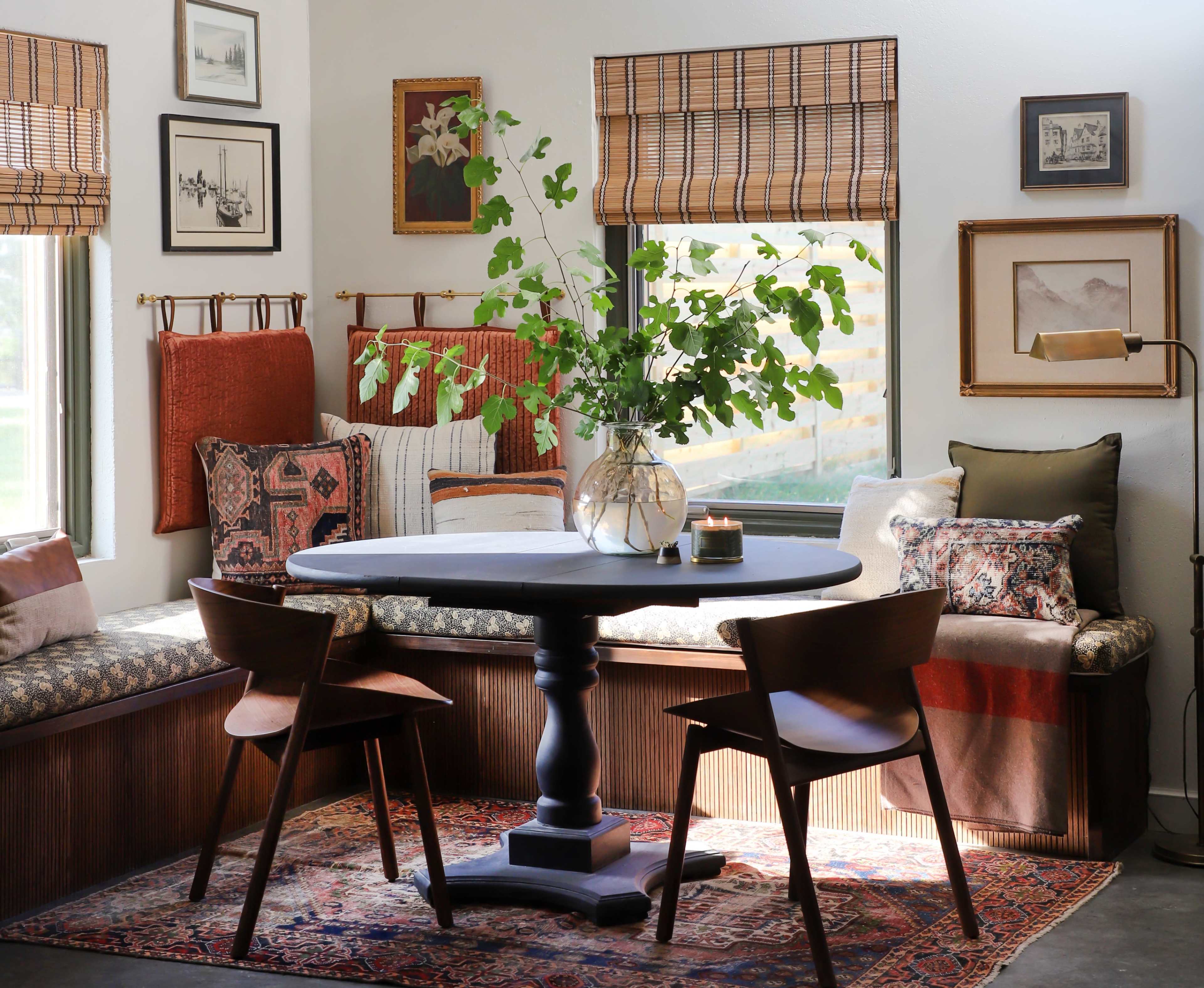 A cozy dining area featuring a round table with two chairs, surrounded by a window seat adorned with patterned pillows and framed artwork on the walls.