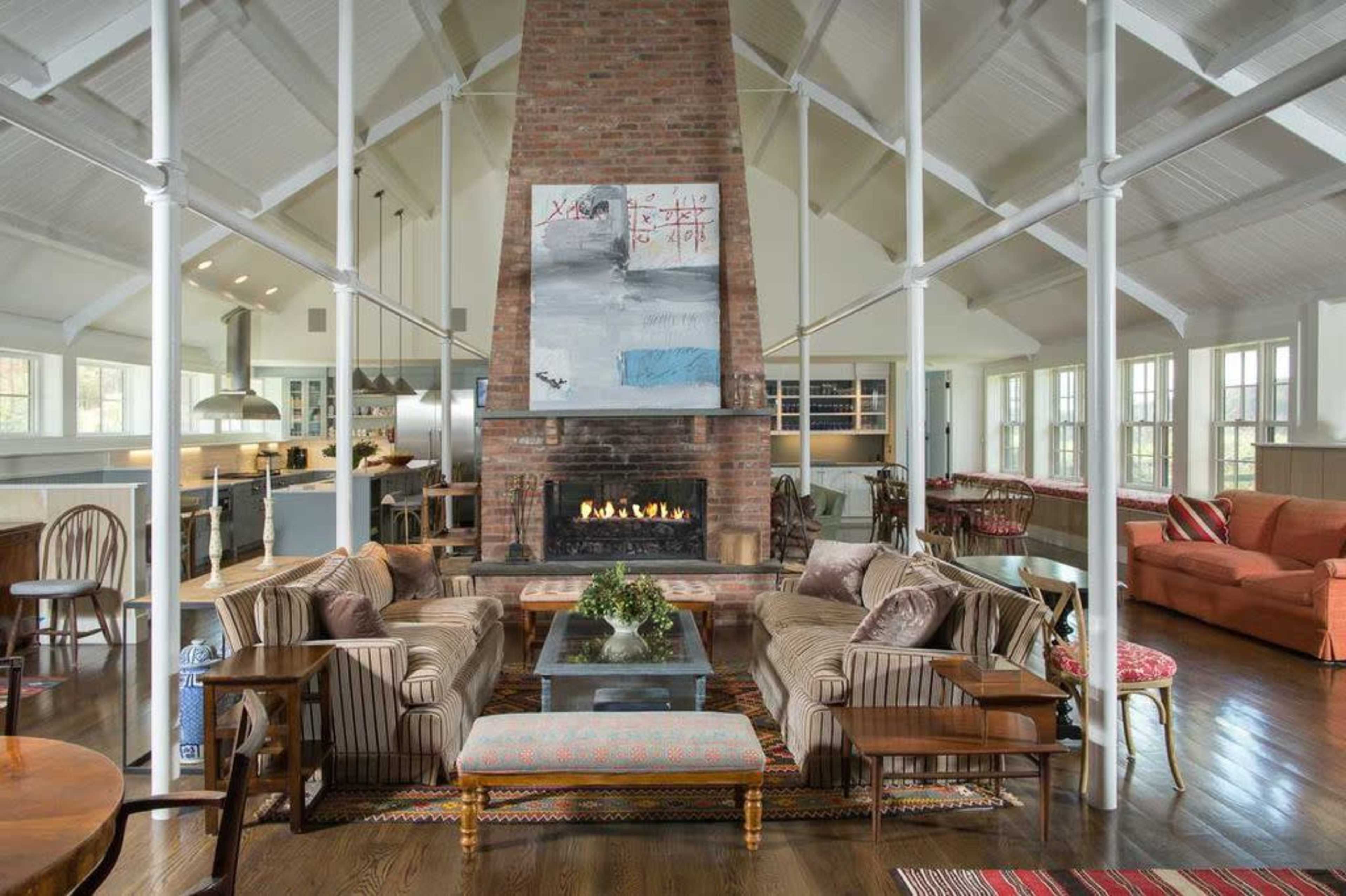 The image shows a spacious living room featuring a central brick fireplace, two sofas, a coffee table, and large windows that provide natural light.