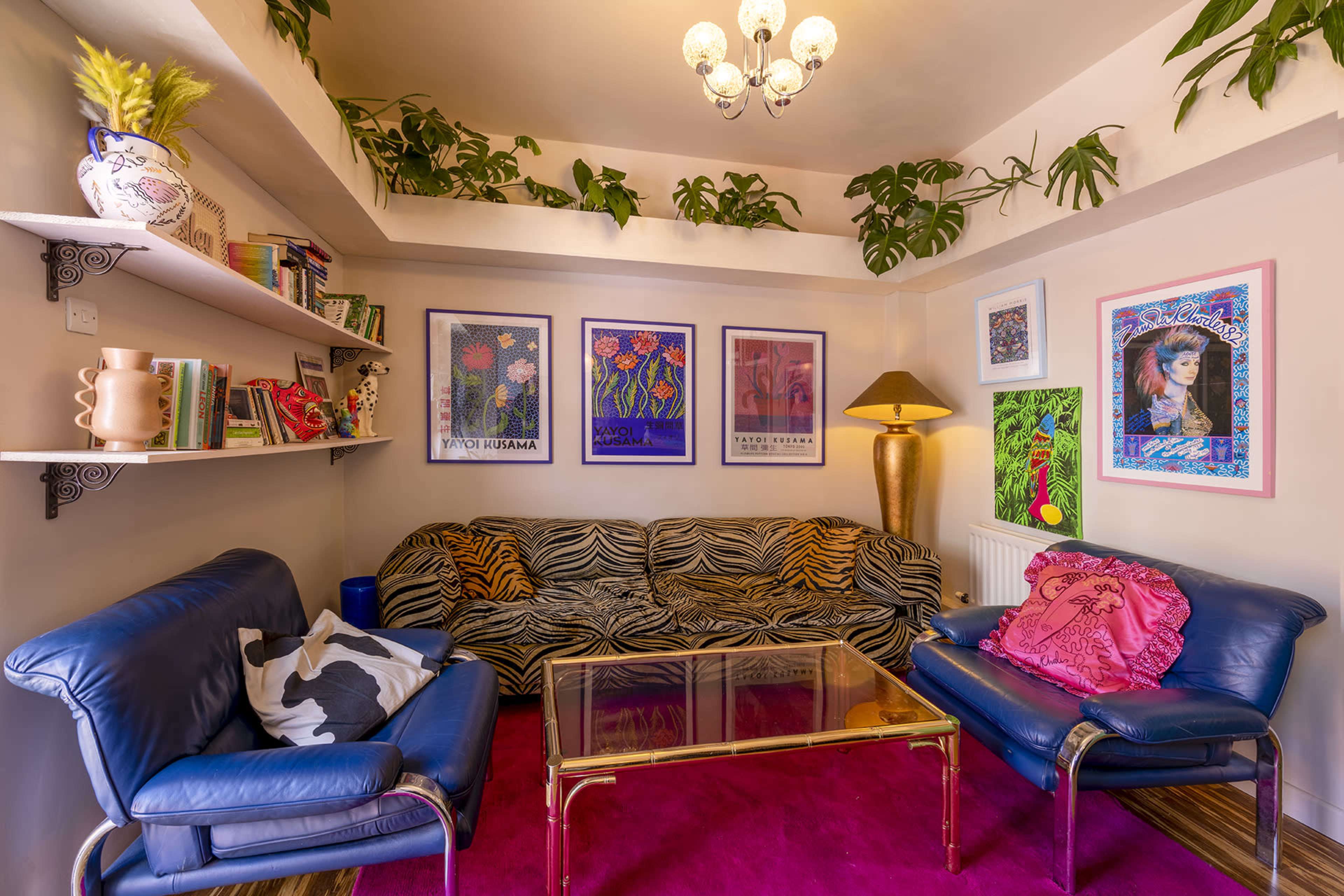 A living room with a zebra-striped couch, blue chairs, a glass coffee table, colorful wall art, potted plants, and a light fixture.