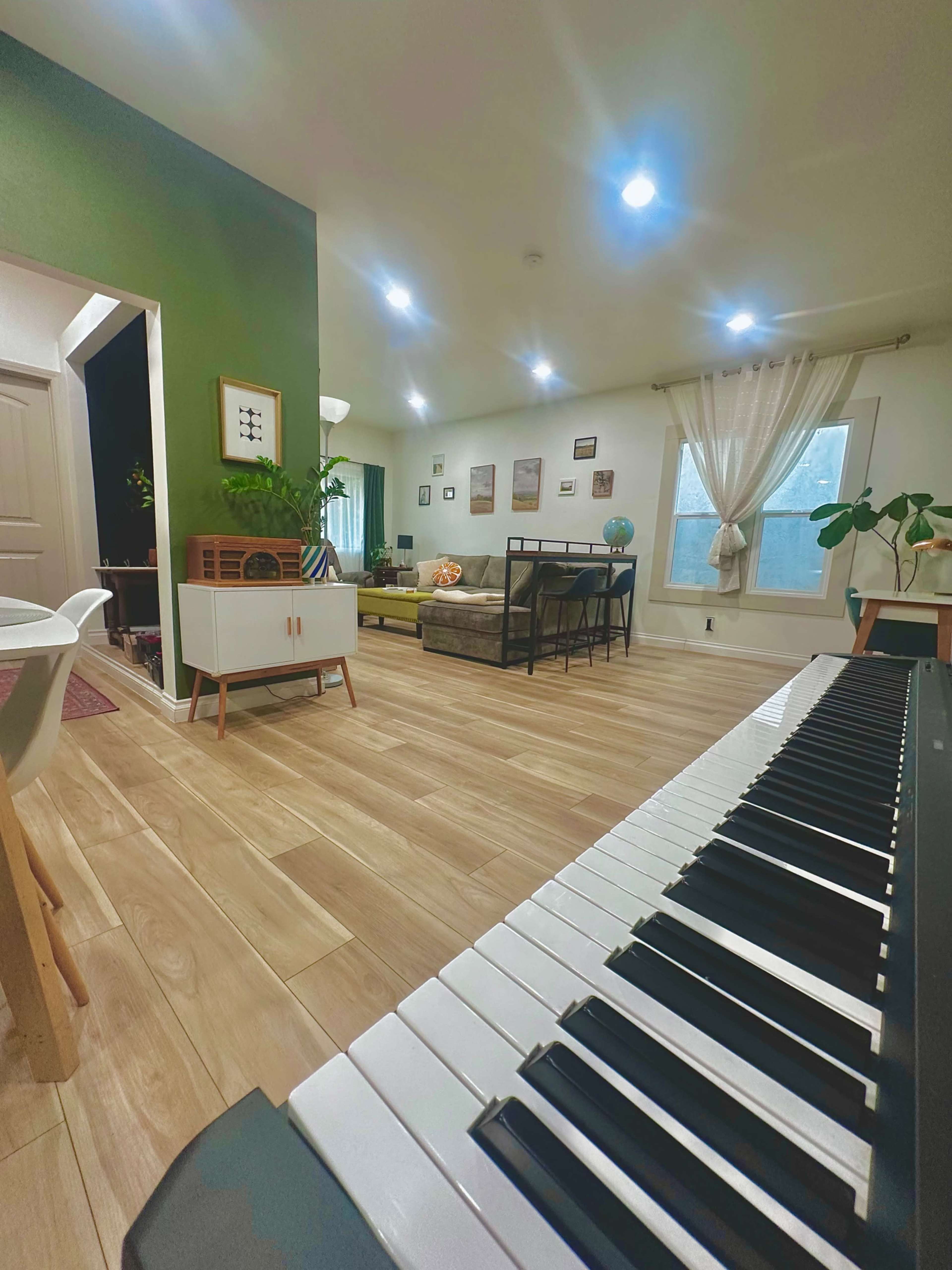 The image shows a spacious living area with wooden flooring, a variety of seating, and natural light coming through large windows.