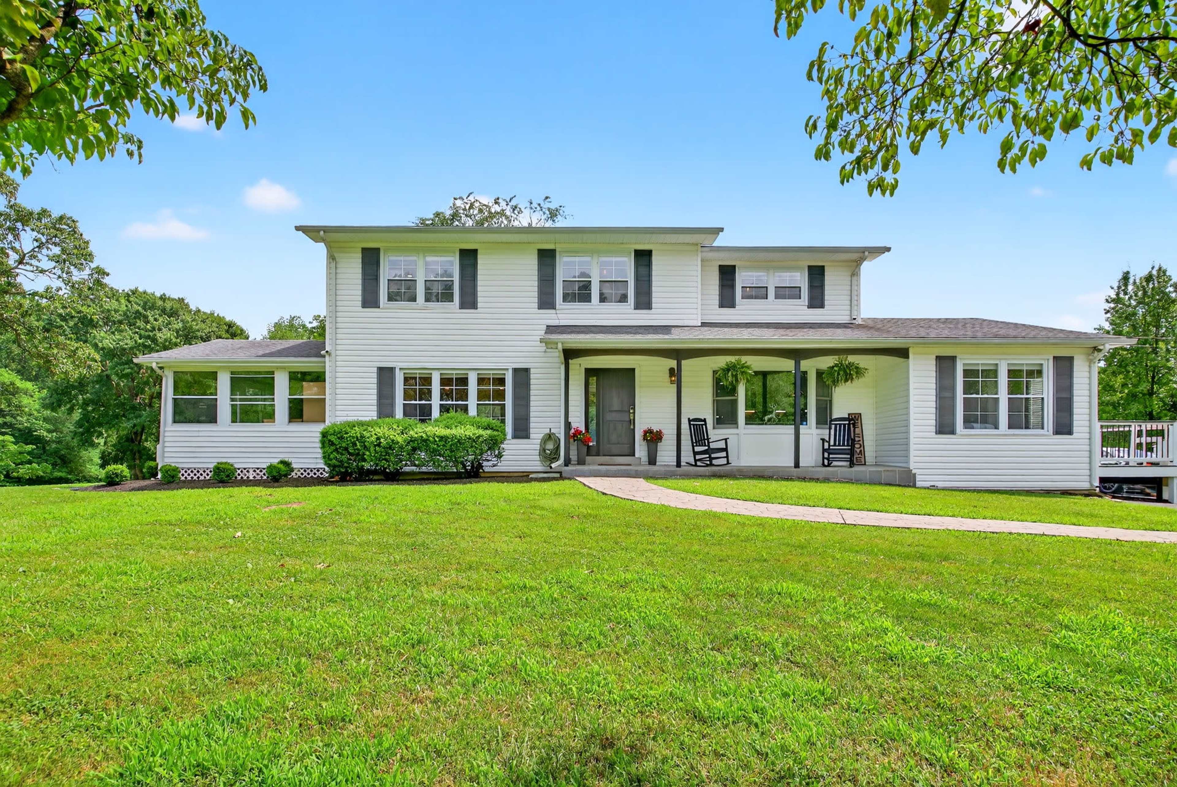 A large, two-story white house with a front porch and rocking chairs is surrounded by a green lawn and trees.