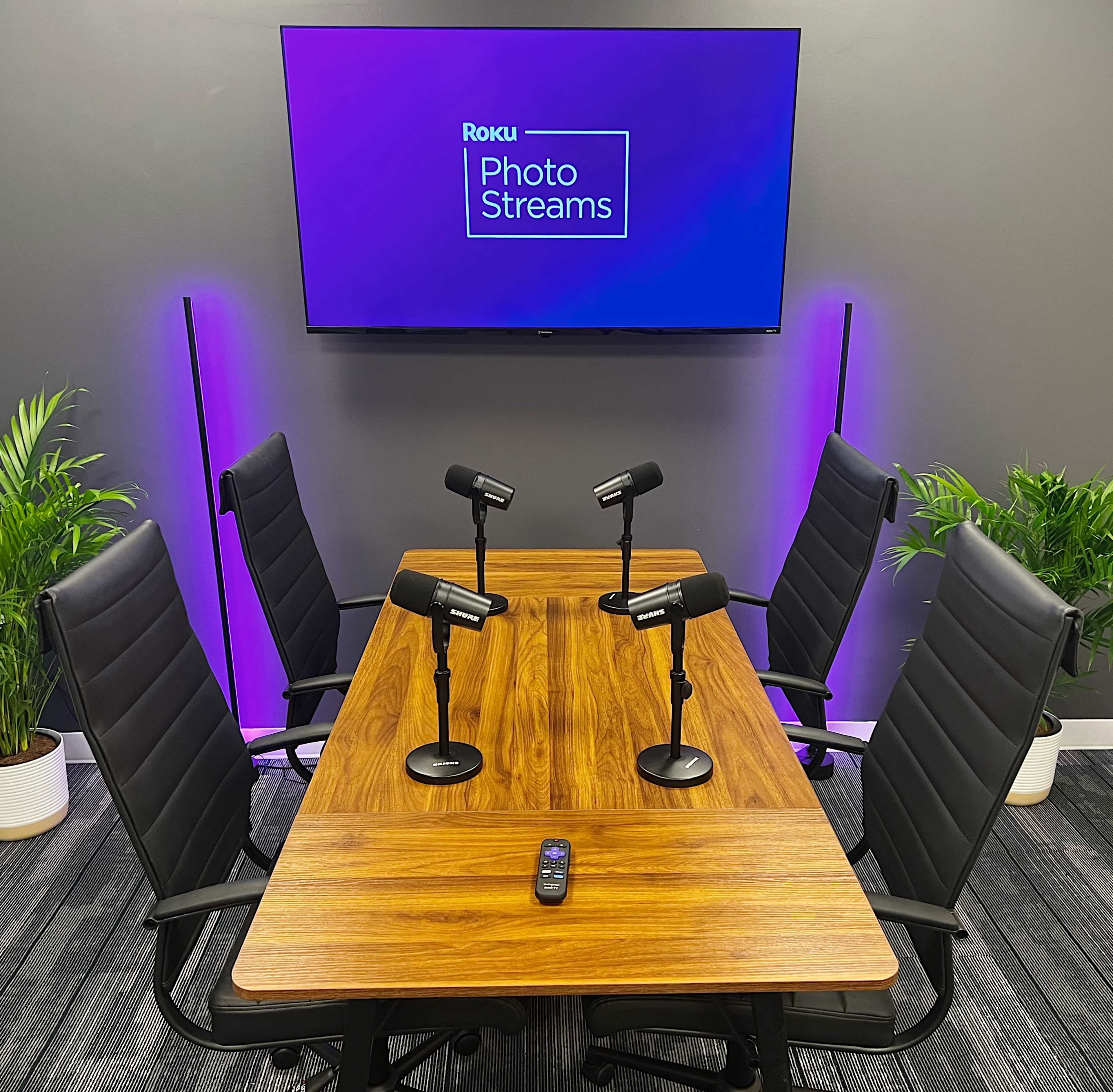 A conference table with four microphones is positioned in front of a wall-mounted screen displaying "Roku Photo Streams."