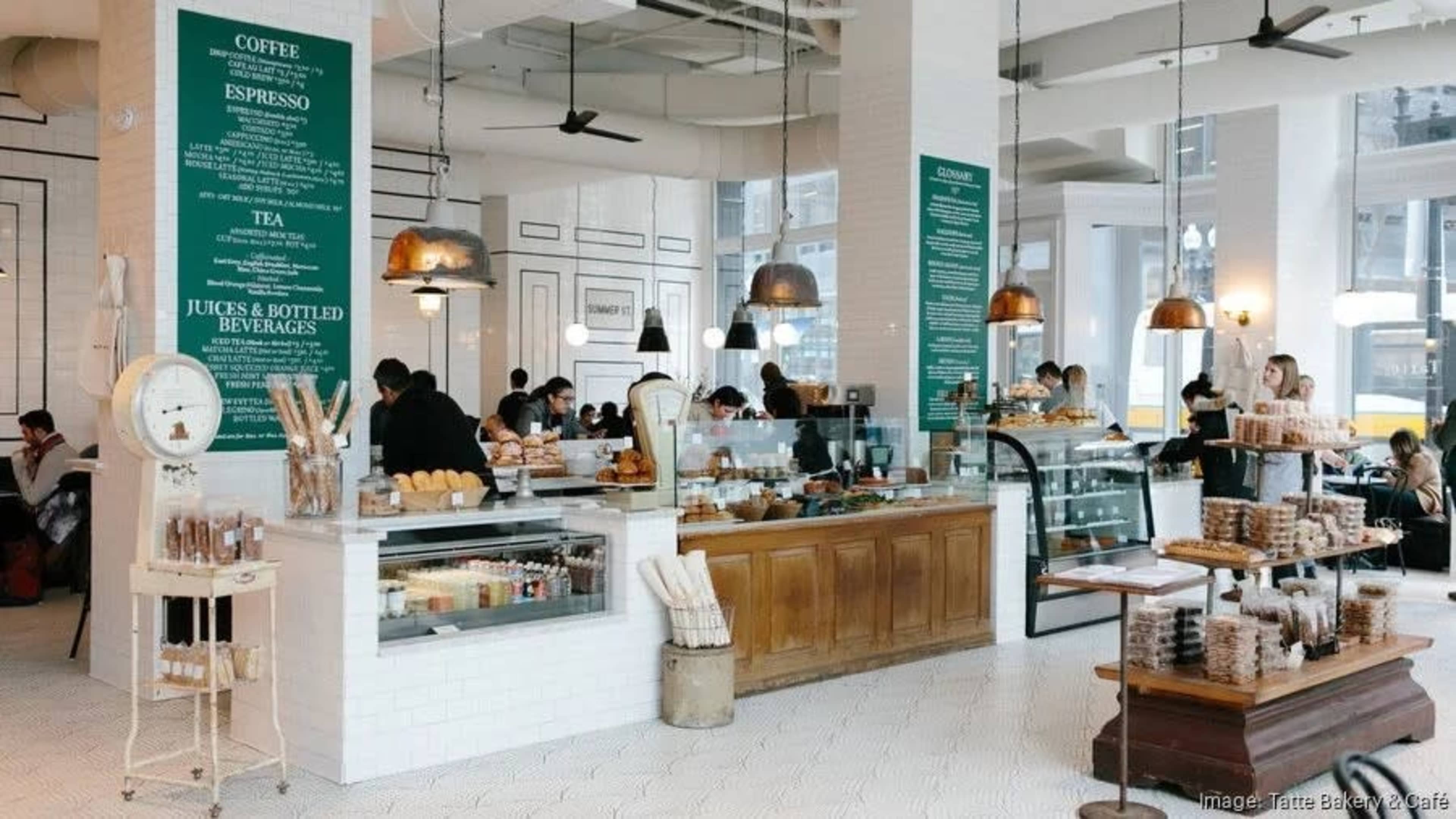 A busy café features a glass display case for pastries and beverages, with customers seated at various tables and a menu displayed on the walls.