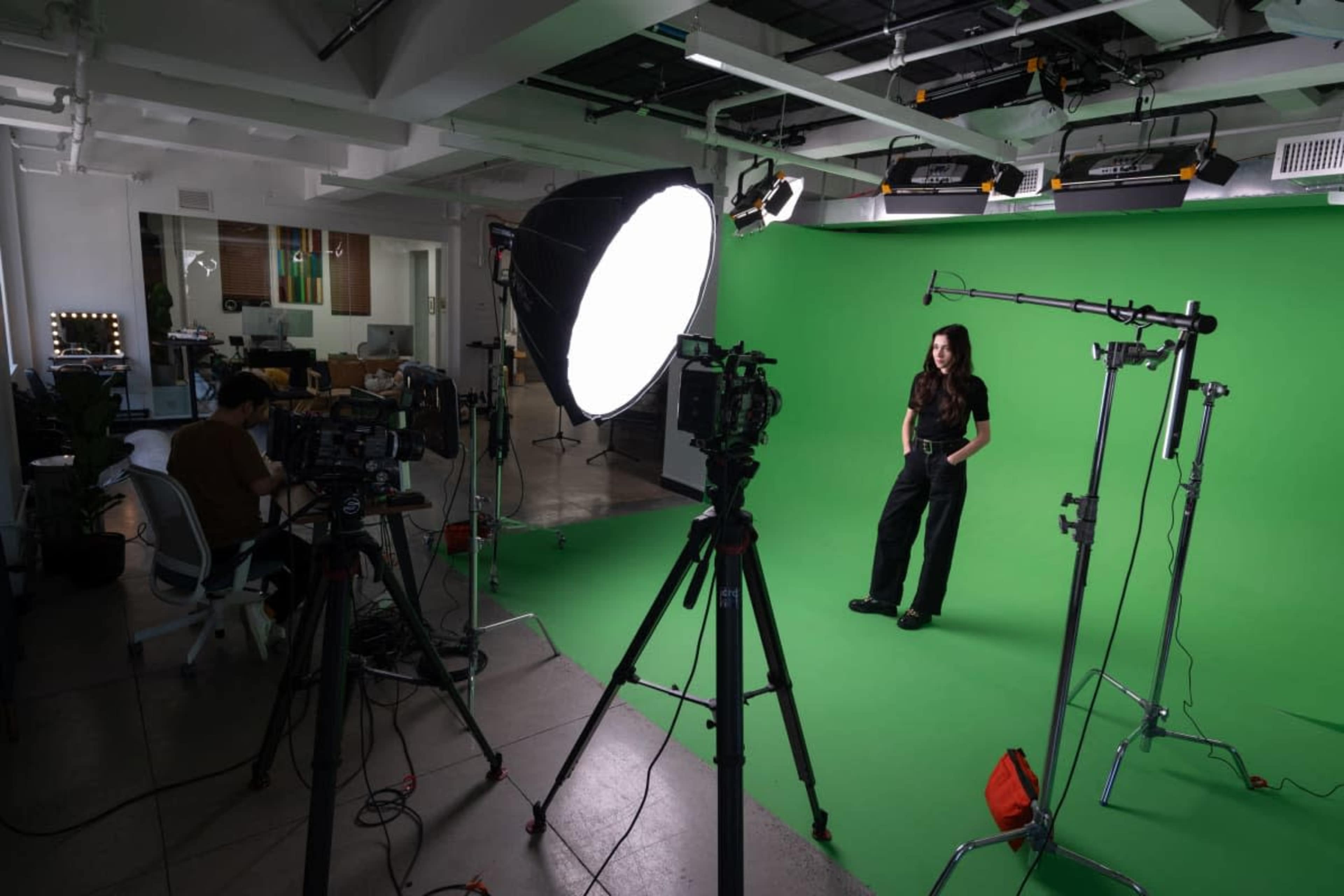 A person stands on a green screen backdrop in a studio, surrounded by cameras and lighting equipment.