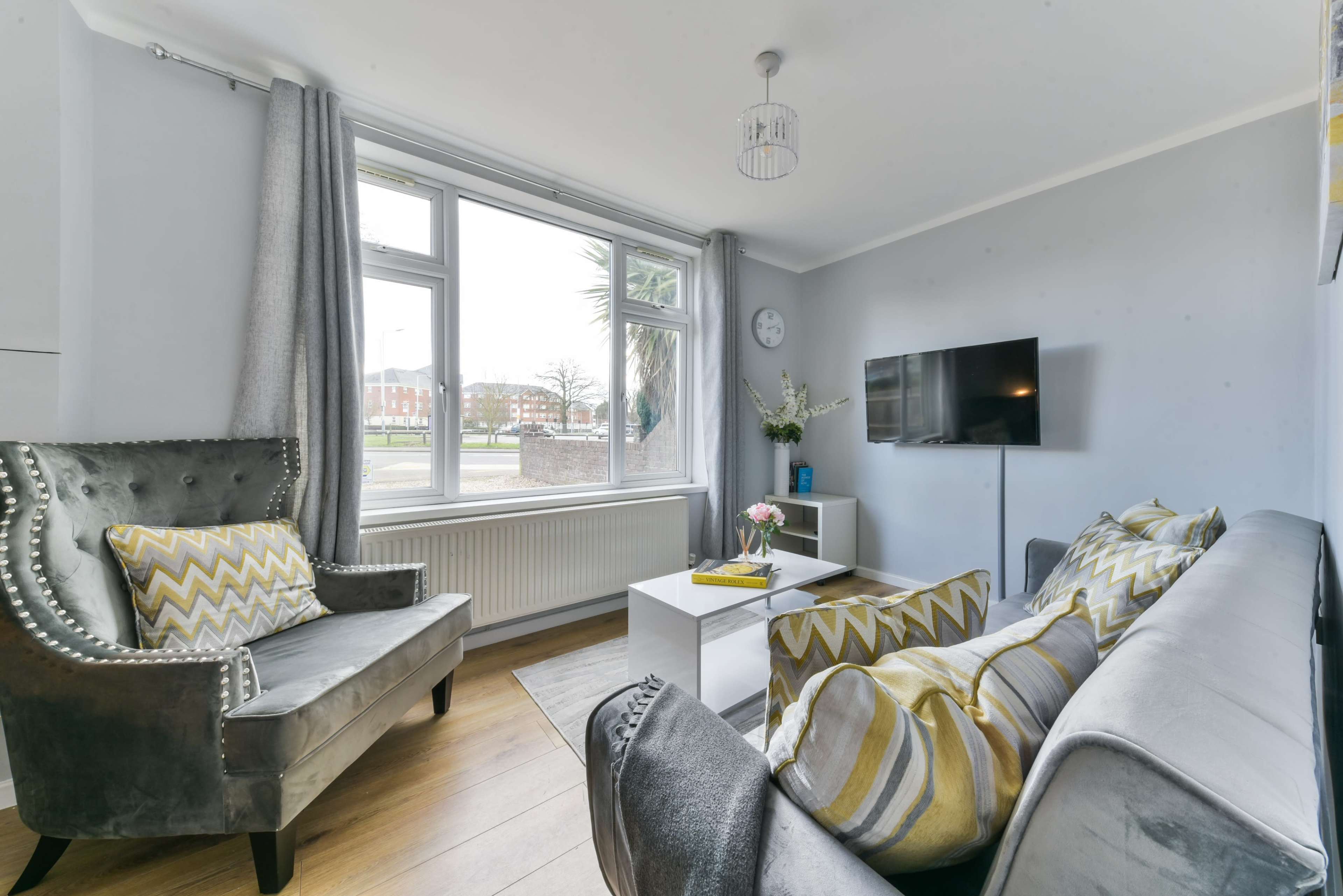 A cozy living room features a gray sofa and armchair, a coffee table with flowers, and a wall-mounted TV near a large window.