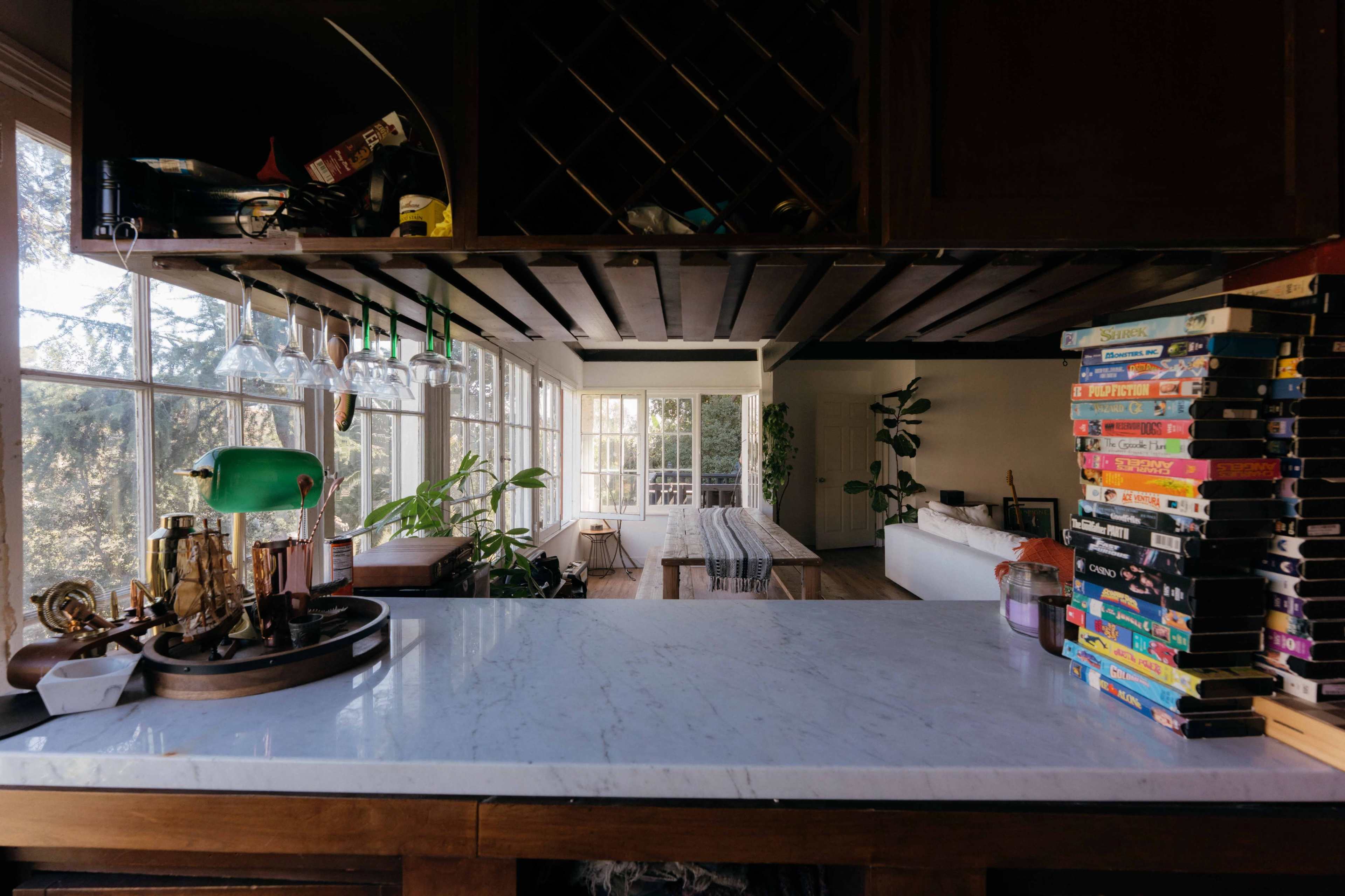 The image shows a kitchen bar with a marble countertop, glassware hanging above, and a stack of VHS tapes on one side, leading into a bright, open living space with large windows and greenery.
