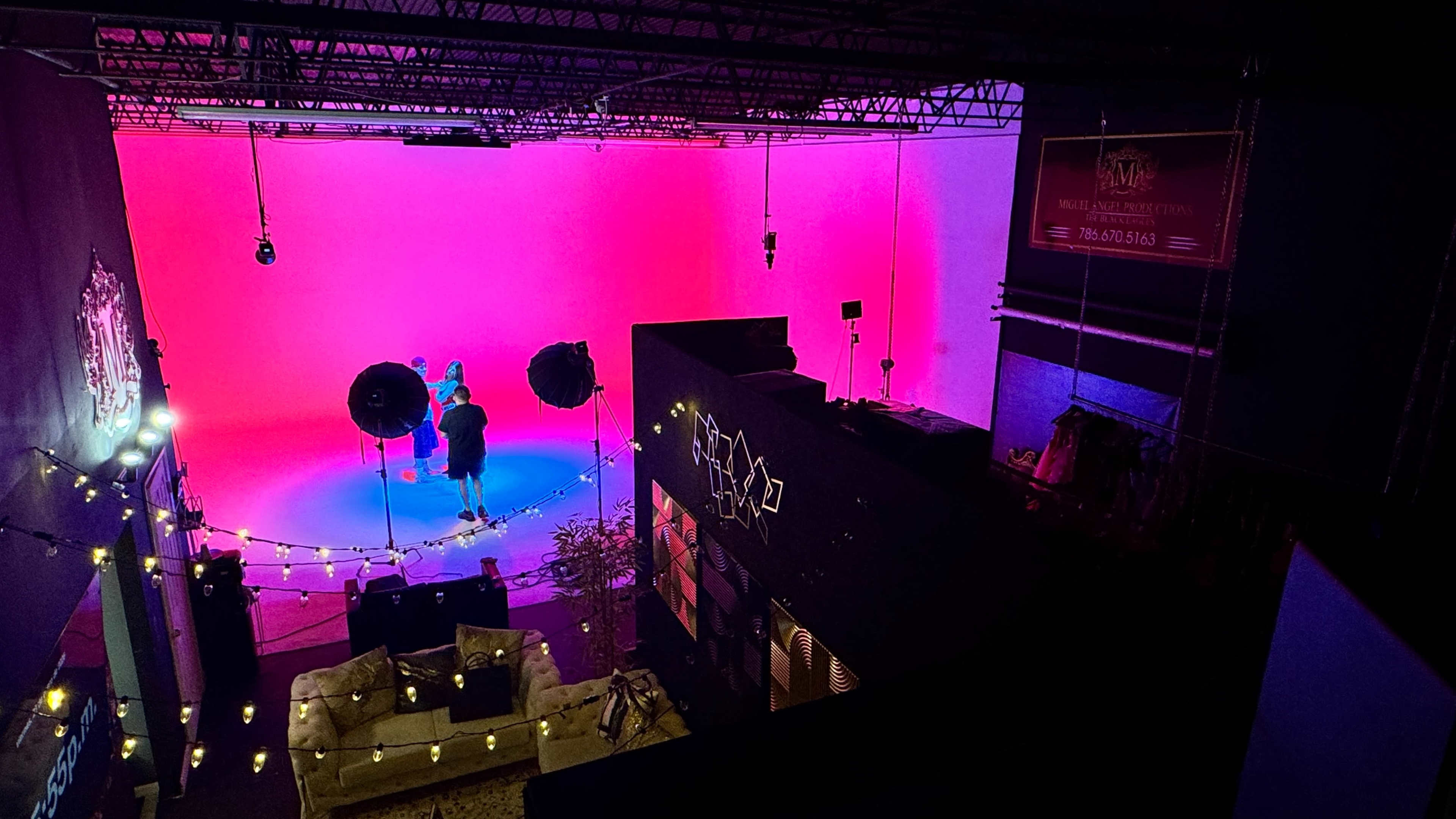 A studio space features two large lights and a performer against a bright pink backdrop, with a cozy seating area visible in the foreground.