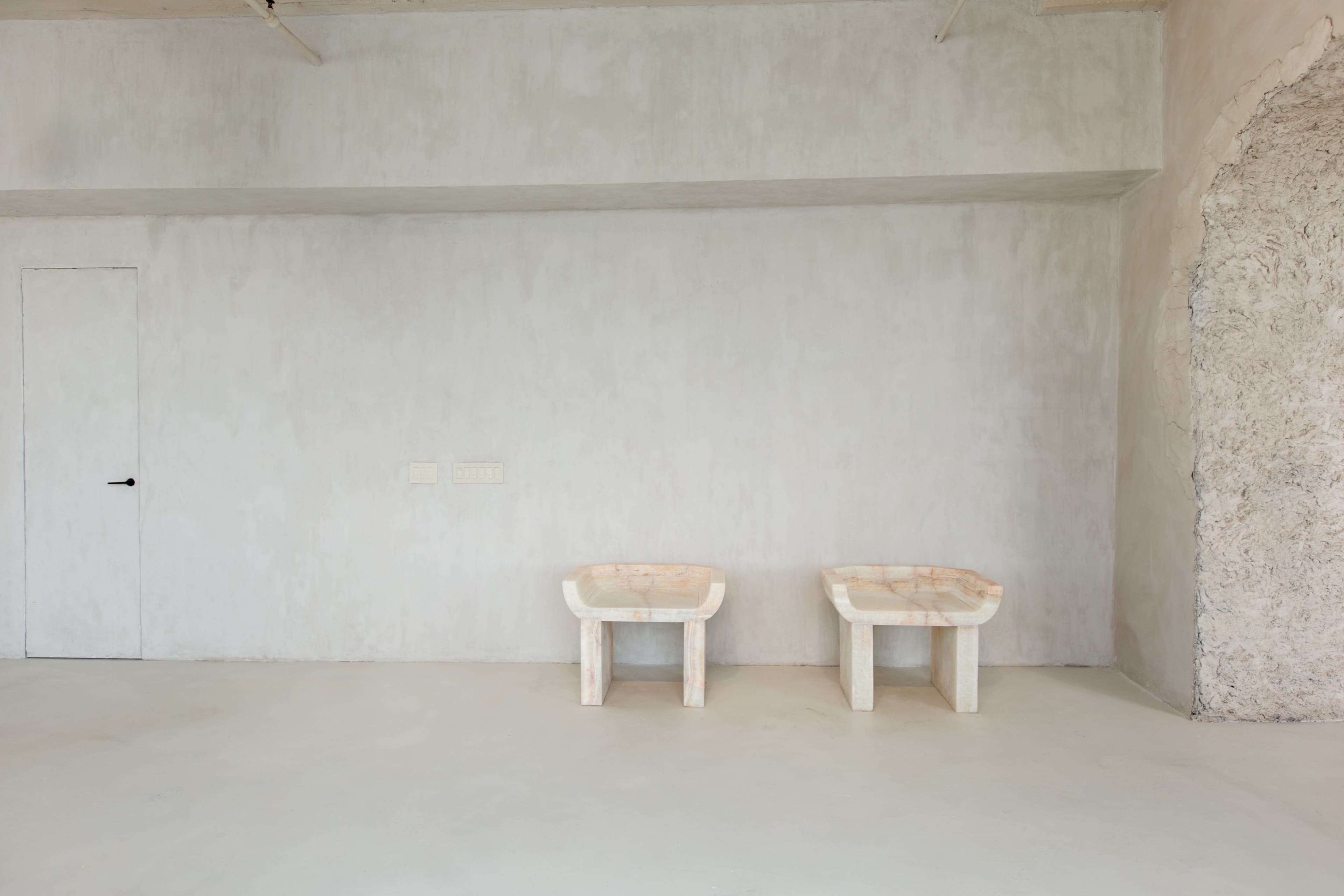 The image shows a minimalist room with a plain gray wall and two simple, marble-patterned chairs placed side by side on a concrete floor.