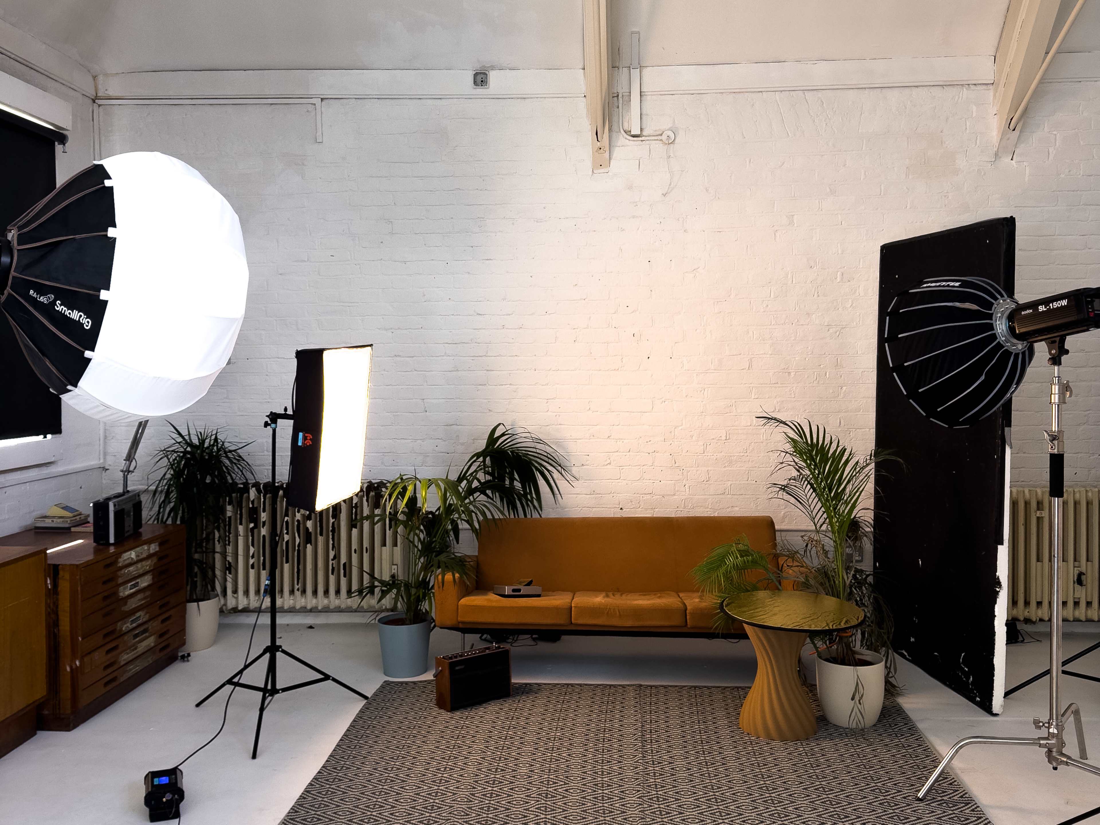 The image shows a well-lit photography studio featuring a brown couch, two large softbox lights, a round table, and various plants against a white brick wall.